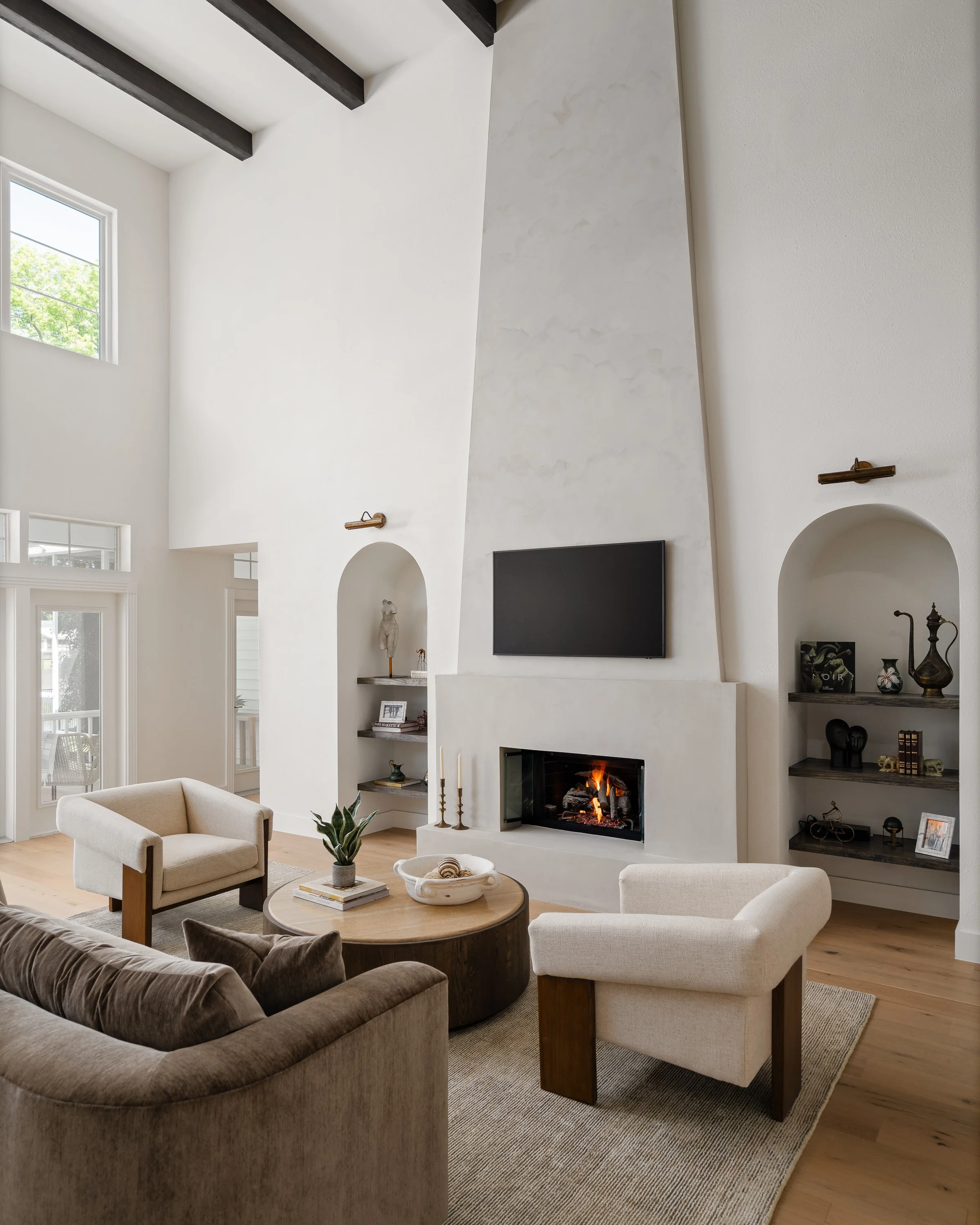 Modern living room with a fireplace, wall-mounted TV, beige and brown furniture, wooden shelves with decorative items, large windows, and high white ceilings with dark exposed beams.