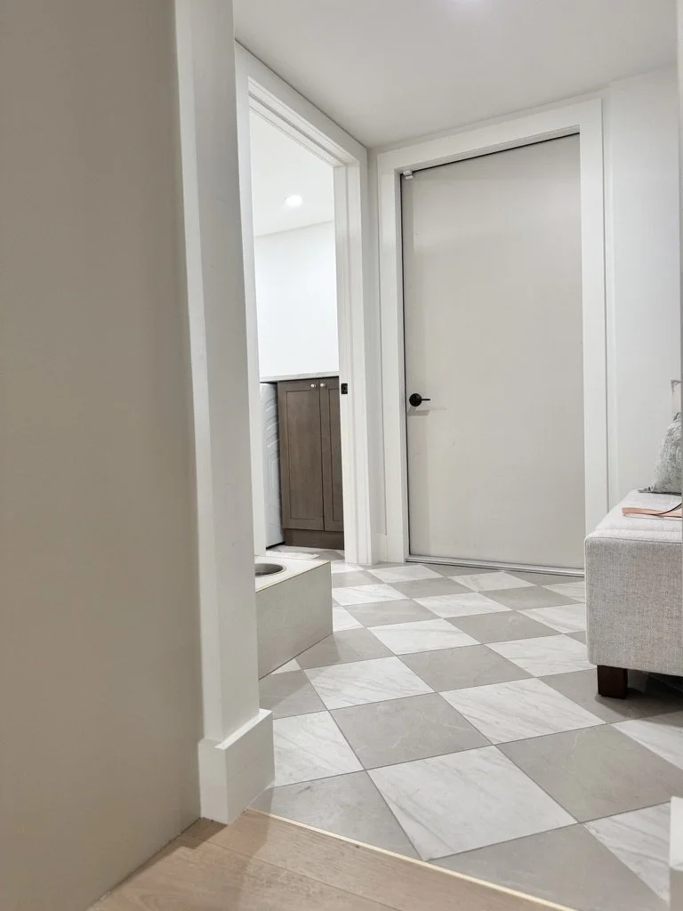 Interior view of a room with checkered gray and white tile floor, white walls, a white door, a small cabinet with a dark wood finish, part of a gray sofa, and a laundry area with a washing machine.