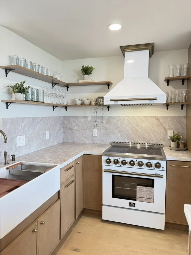 Kitchen with beige cabinets, marble countertops, open wooden shelves with glassware and decor, white range hood, and a white oven stove.