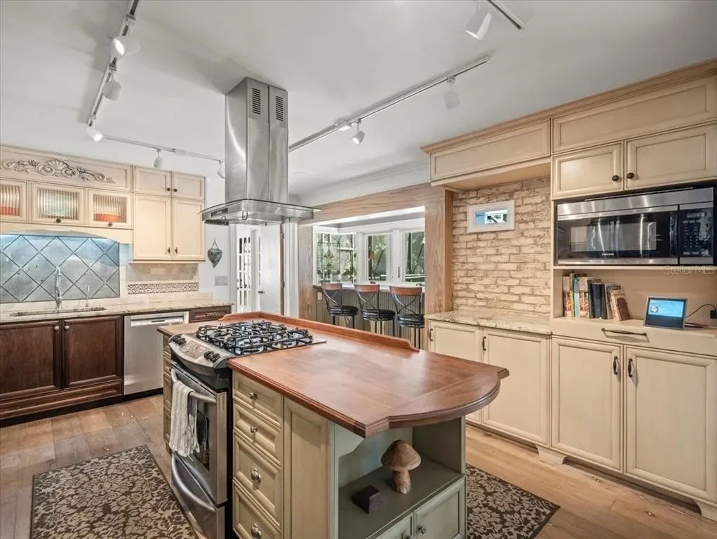 Kitchen with light-colored cabinets, a wooden island, a stainless steel range with a vent hood, bar stools by a window, and a brick accent wall.