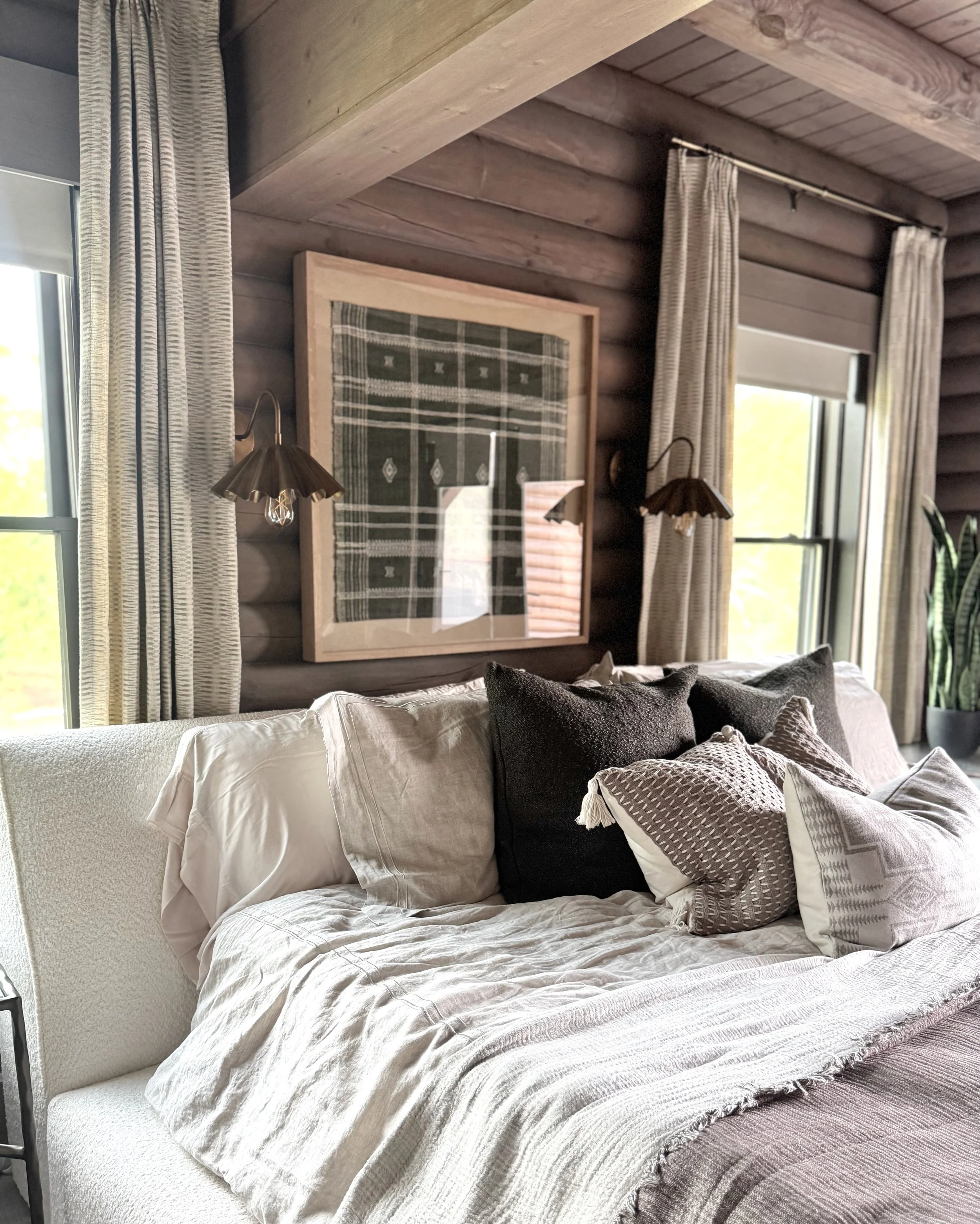 A cozy bedroom with a beige bed covered in multiple pillows, dark and light throw blankets, flanked by two windows with beige curtains. A framed textile artwork hangs on a wooden wall, with two wall-mounted lamps on either side.