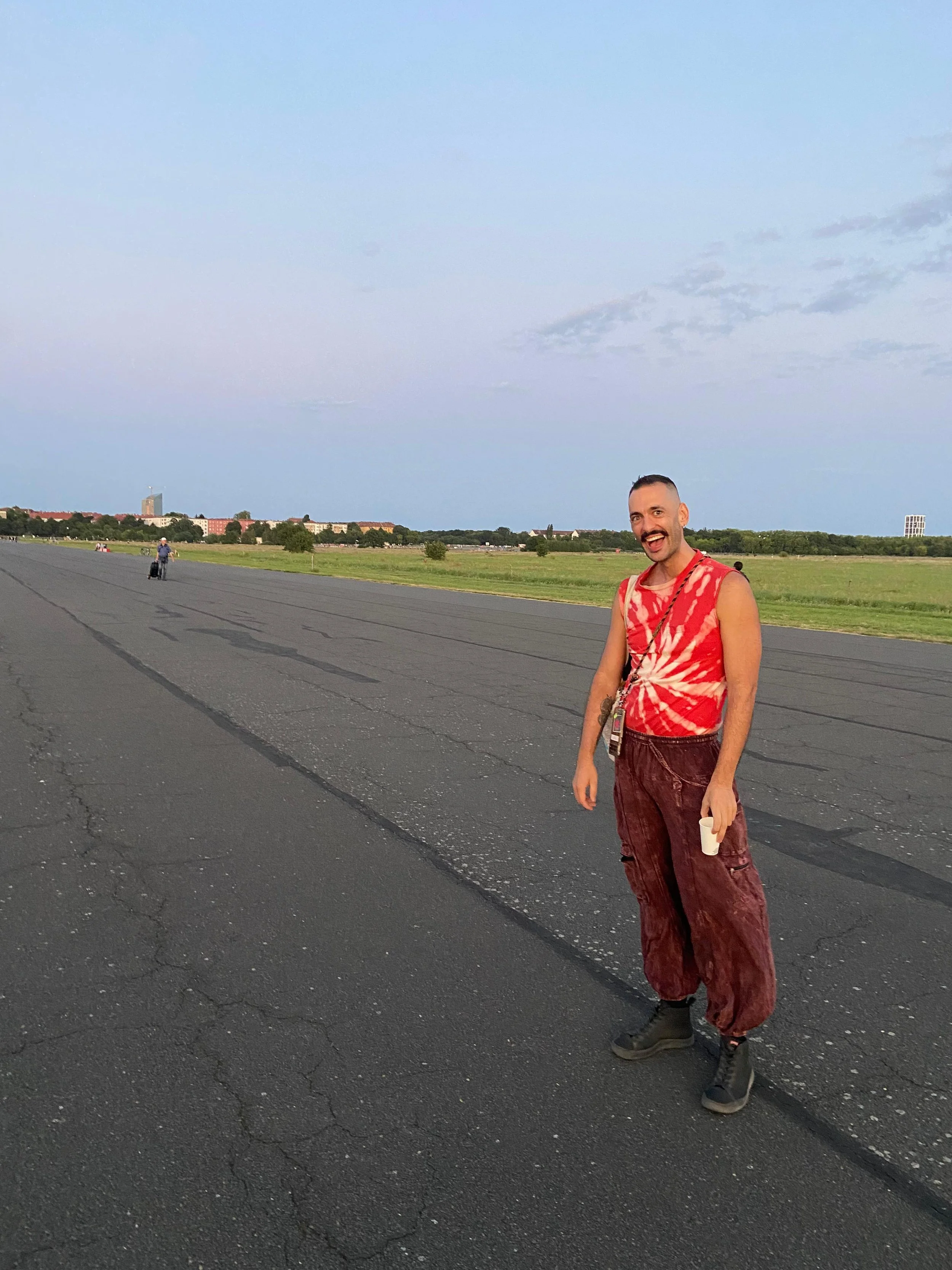 A person standing on a paved road outdoors, wearing a sleeveless red and pink tie-dye shirt, red patterned pants, black boots, and holding a cup, with a landscape of green fields and a cityscape in the background under a partly cloudy sky.