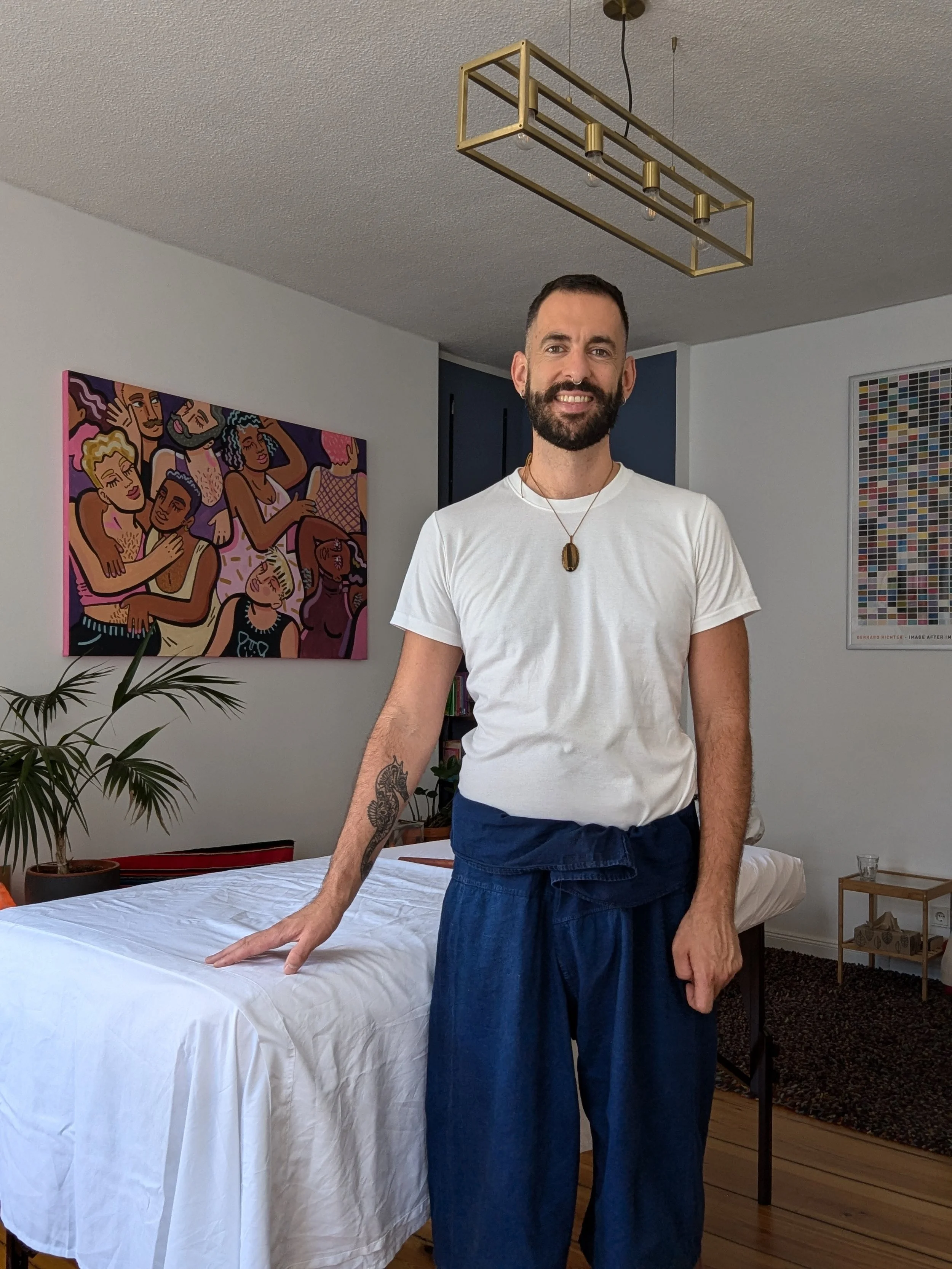 A man with a beard and short dark hair wearing a white T-shirt, dark blue pants, and a necklace, standing in a room next to a table with a white cloth, in front of a wall with colorful artwork, and a gold geometric light fixture hanging from the ceiling.