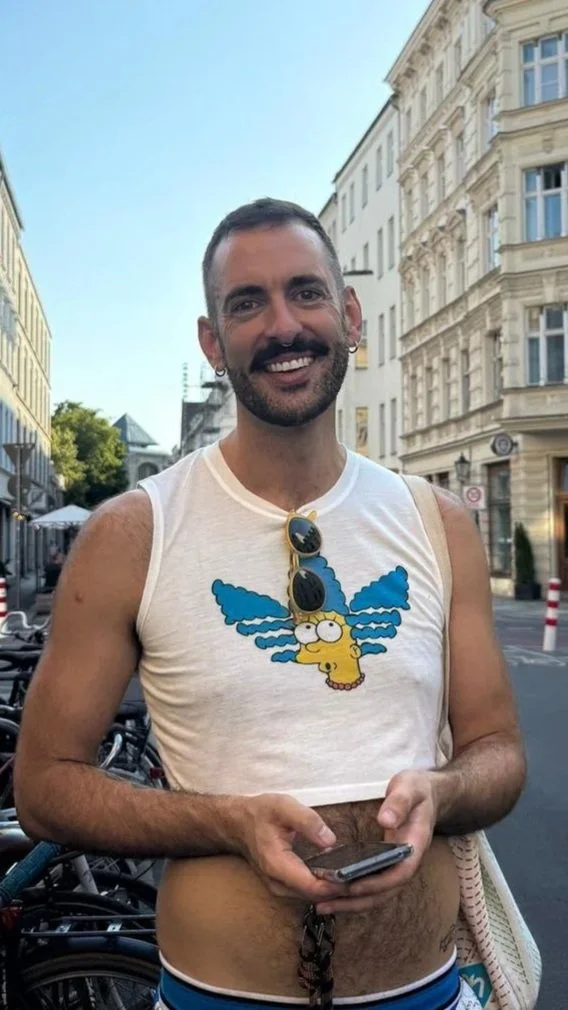 A smiling man with short dark hair and a beard, wearing sunglasses, a sleeveless white shirt with a cartoon character, and holding a phone, standing on a city street with historic buildings and bicycles in the background.