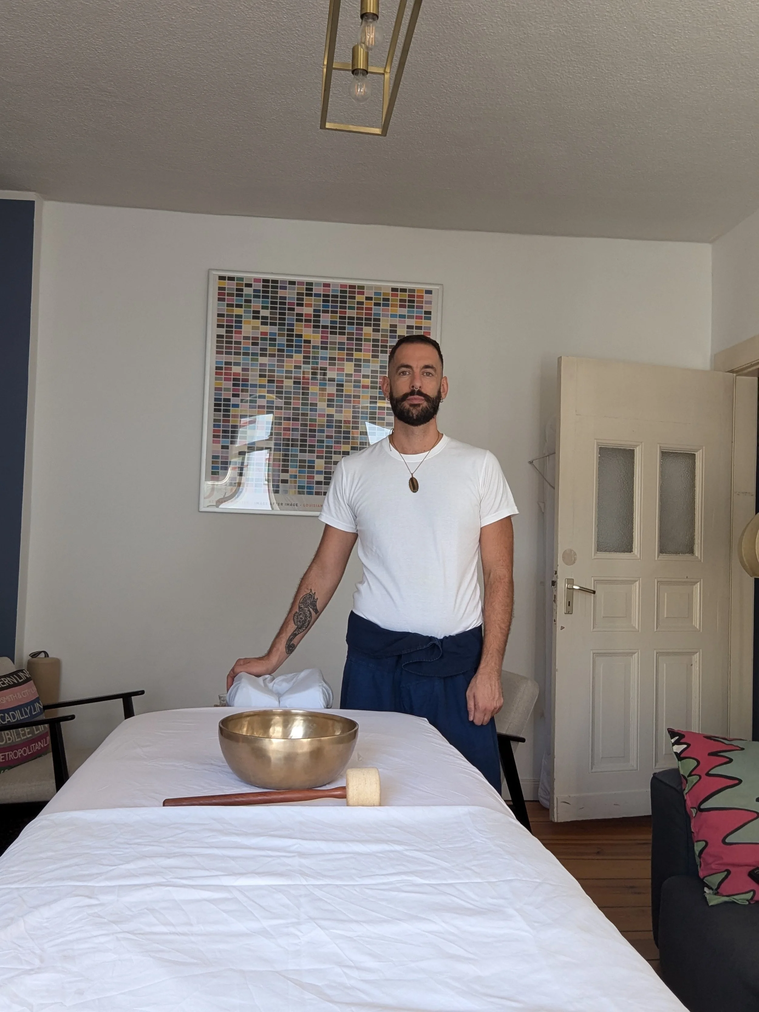 A man with a beard and tattoos on his arm standing beside a table in a room with white walls and a colorful framed artwork. Objects on the table include a brass bowl, a wooden mallet, and a white cloth. There is a door slightly open, a black armchair with a patterned pillow, and chairs around the table.