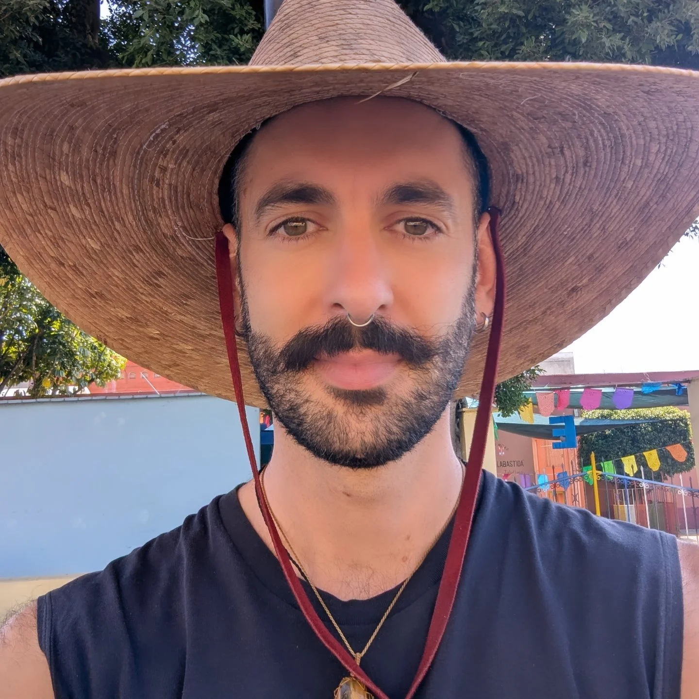 A man with a beard, wearing a large straw hat, black sleeveless shirt, and multiple necklaces, standing outdoors with colorful decorations in the background.