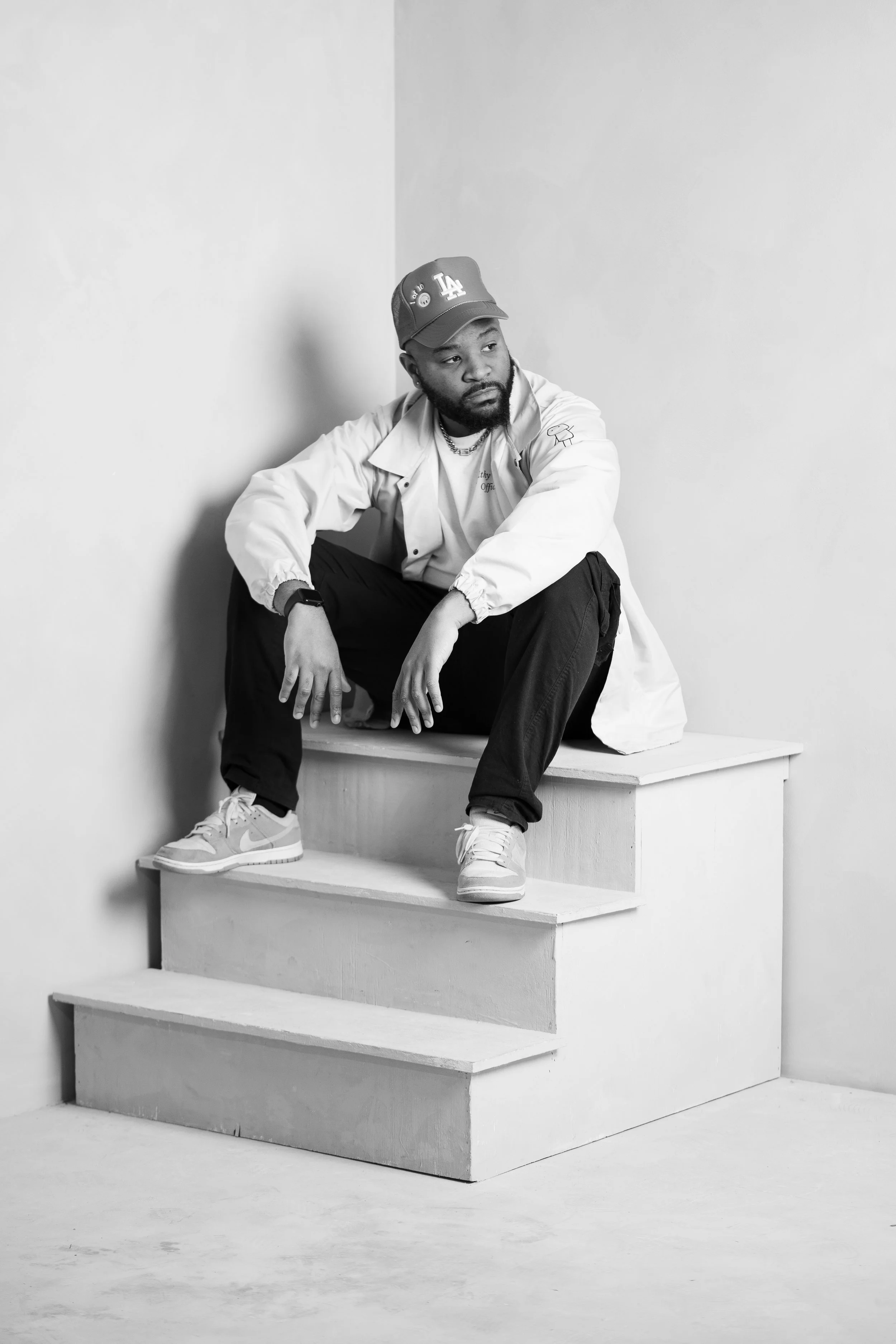A Black man wearing a LA Dodgers baseball cap, a light-colored jacket, black pants, and sneakers, sitting on a white wooden platform against a plain light background. He is looking to the side with a contemplative expression.