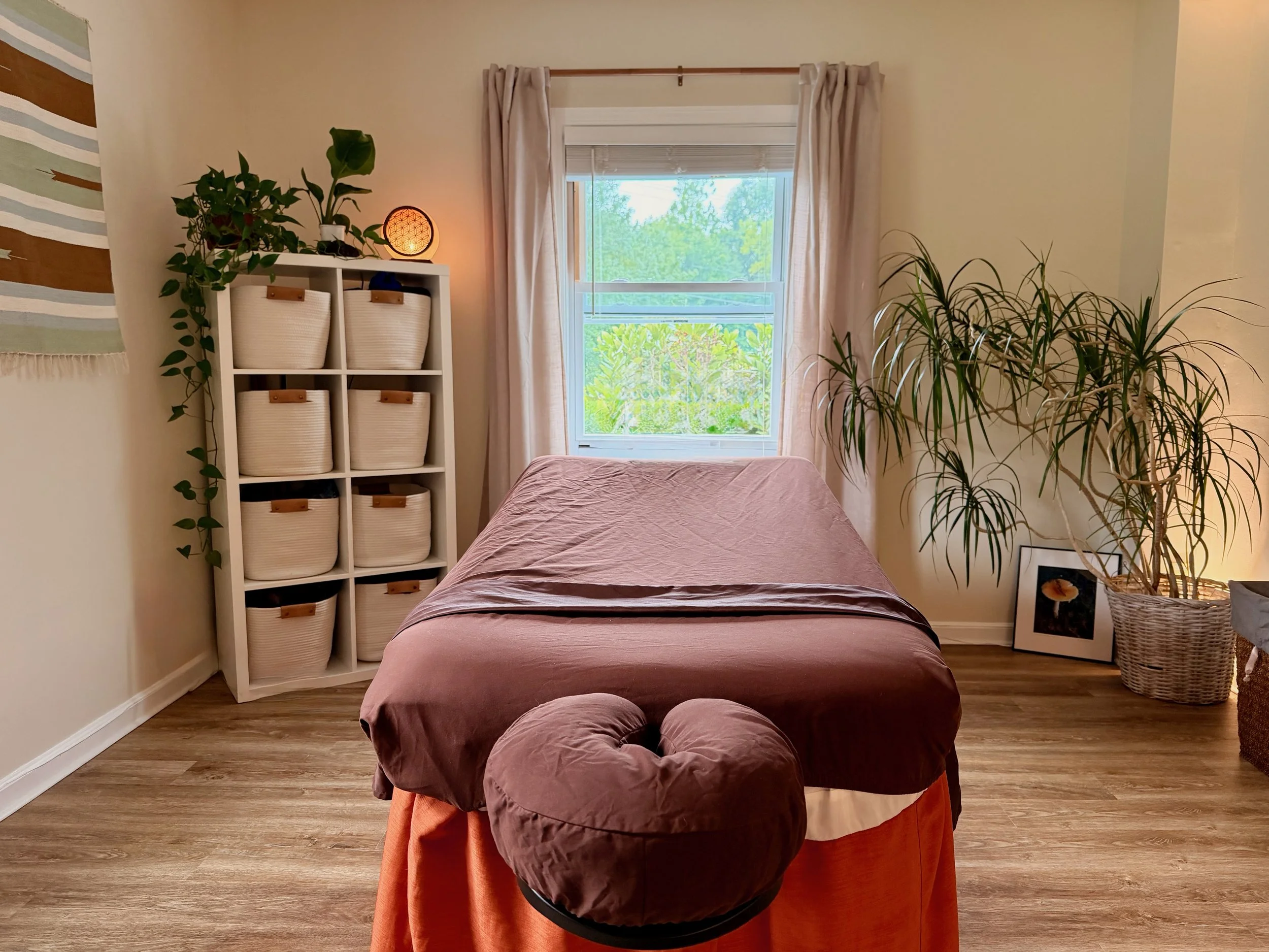 A cozy bedroom with a bed covered in a soft mauve duvet, a small round ottoman at the foot of the bed in matching fabric, a white wooden shelf with baskets and plants, a window with open blinds showing green trees outside, beige curtains, a large houseplant in a basket, and wall art.