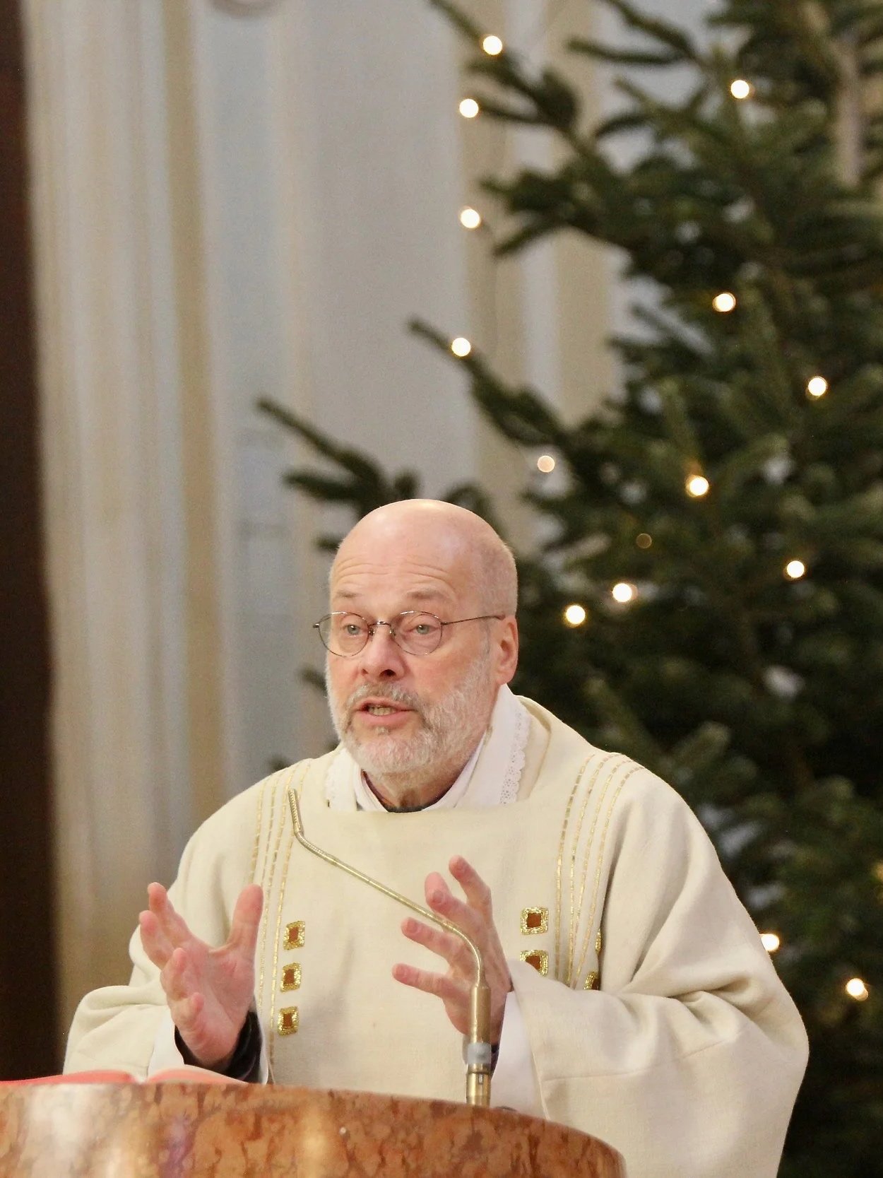 Pfarrer Bernhard Messer spricht während der Messe in der Schubertkirche. Im Hintergrund ein festlich geschmückter Weihnachtsbaum mit Lichtern.