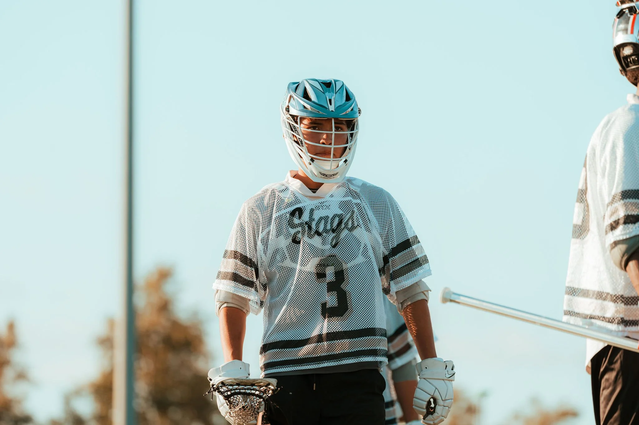A young male lacrosse player wearing a helmet, jersey, and gloves, standing on the field with a lacrosse stick in hand, looking at another player.
