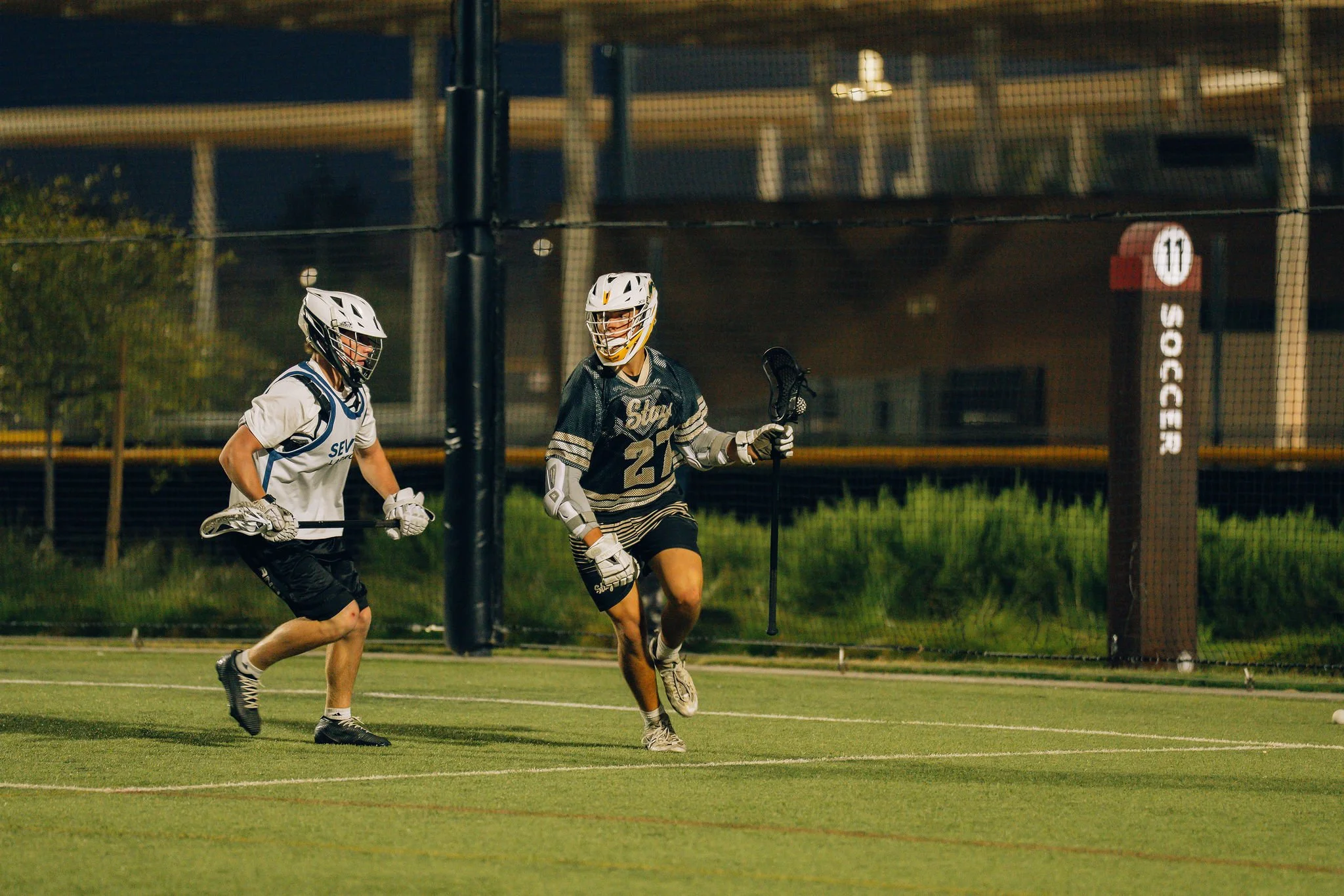 Two male lacrosse players wearing helmets and sports gear are playing on a field at night. One player is in black and white while the other is in white and blue. They are running towards the goal post, which is in the background, under a night sky.