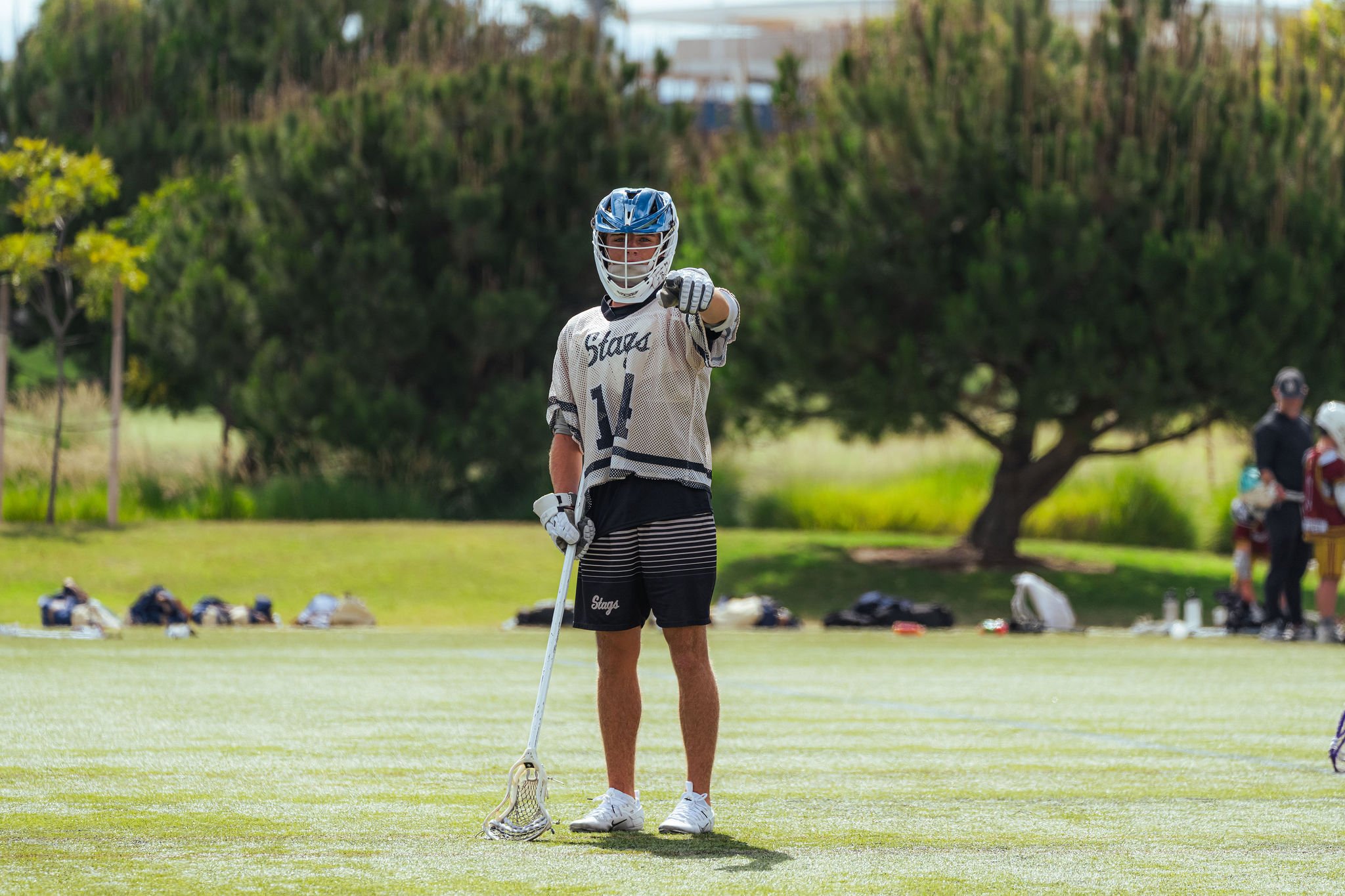 A lacrosse player wearing a blue helmet, white jersey with the number 14, and black shorts stands on a field holding a lacrosse stick, pointing toward the camera. There are trees and other players in the background.