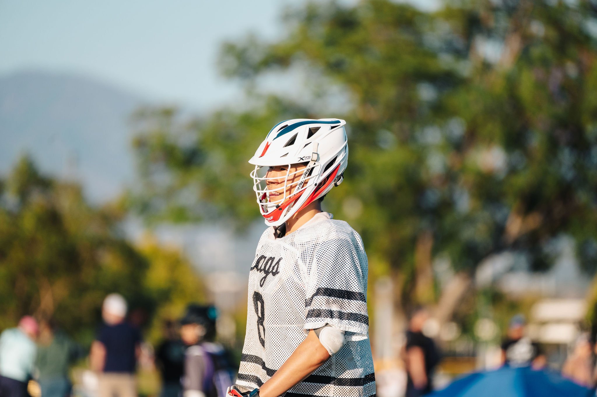 A person wearing a white helmet and sports jersey outdoors, with blurred people and trees in the background.