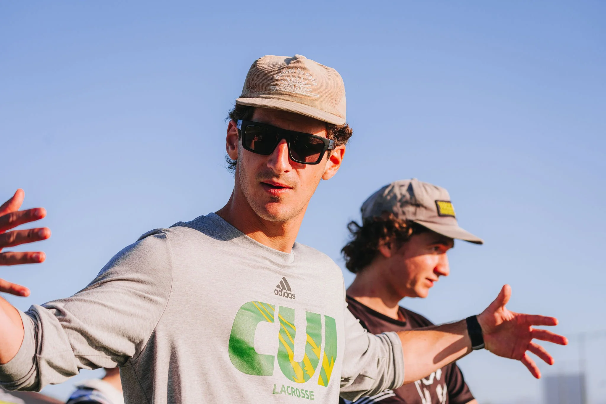 Two young men, wearing hats and sunglasses, are standing outdoors against a clear blue sky, with their arms extended to the sides.