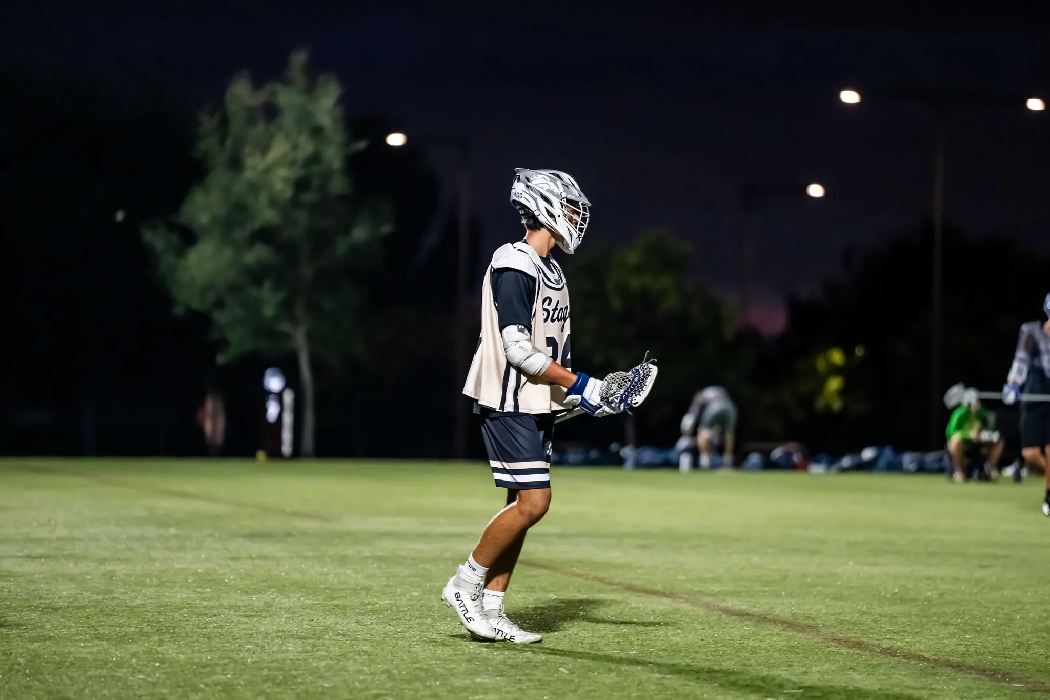 A lacrosse player stands on a field at night, wearing a helmet and sports gear, holding a lacrosse stick, with others visible in the background.