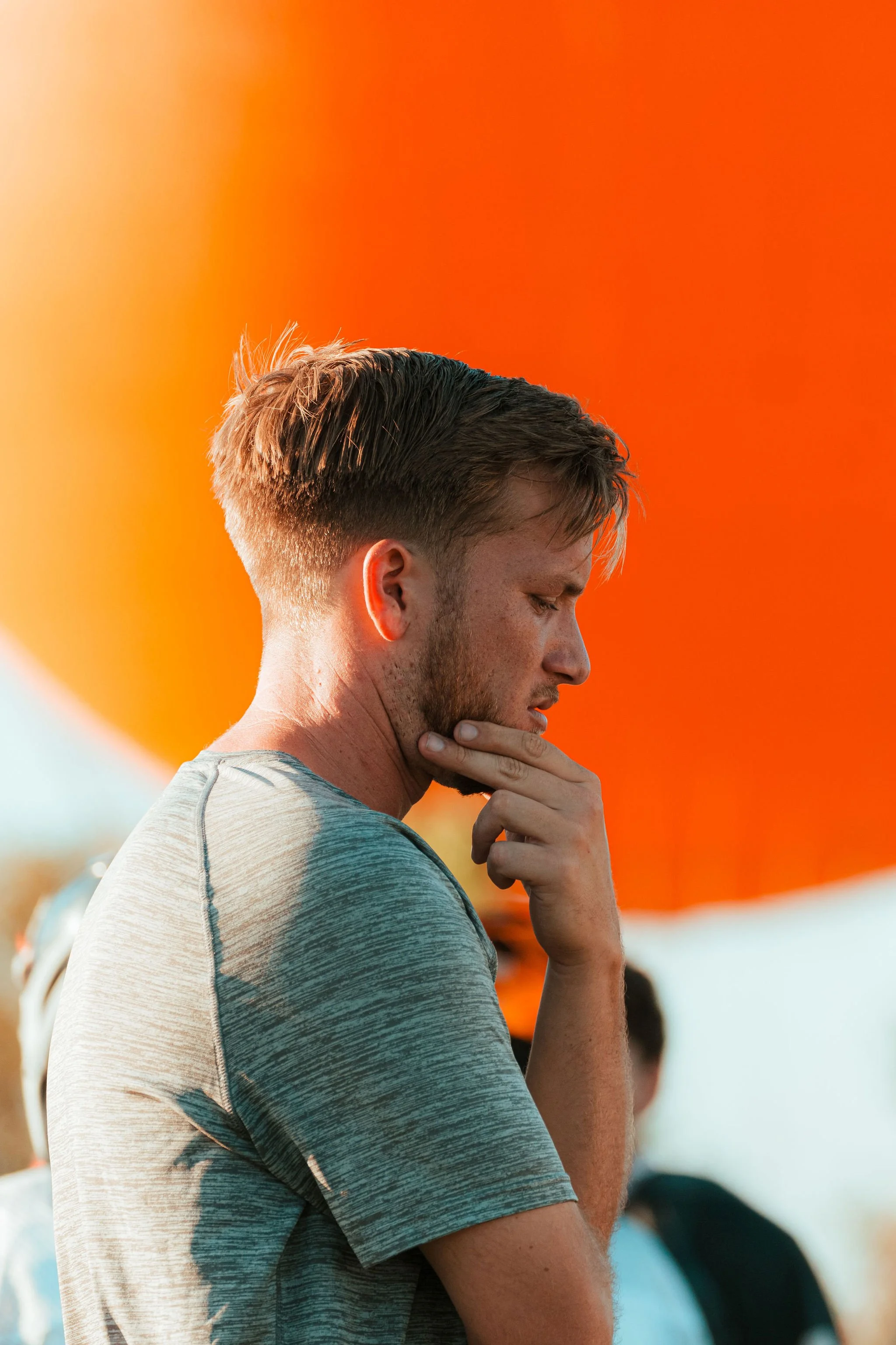 Side profile of a young man with brown hair and a beard, wearing a grey t-shirt, at sunset with an orange sky in the background.