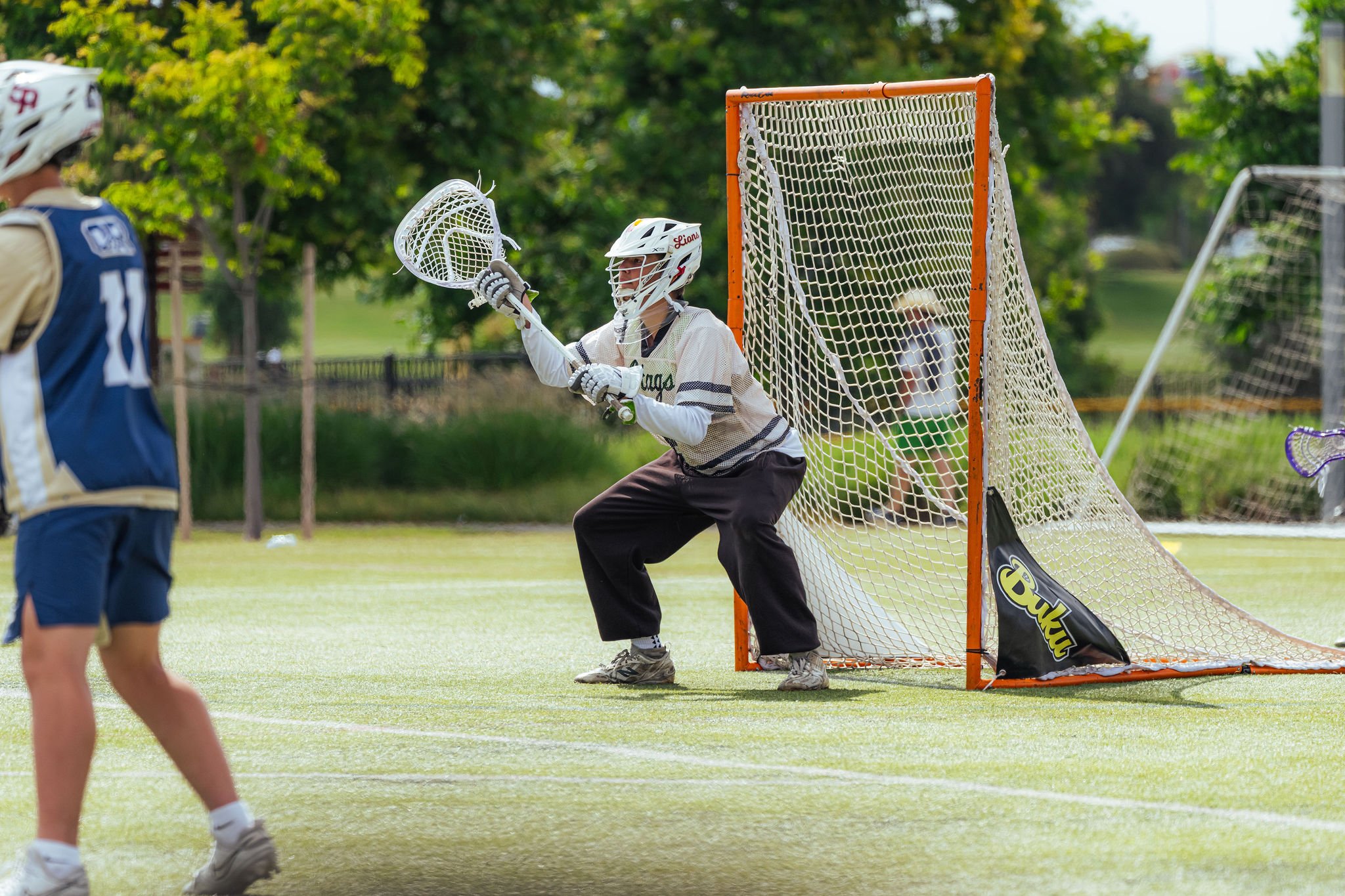 A youth playing lacrosse, holding a lacrosse stick near the goal, with a goalie preparing to block a shot during a game outdoors on a grassy field with trees in the background.
