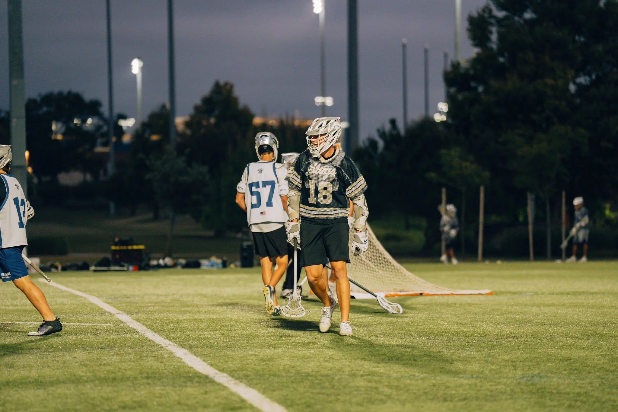 Lacrosse players on a field at dusk, some wearing helmets and jerseys, preparing for practice or a game, with a goal net on the ground nearby.