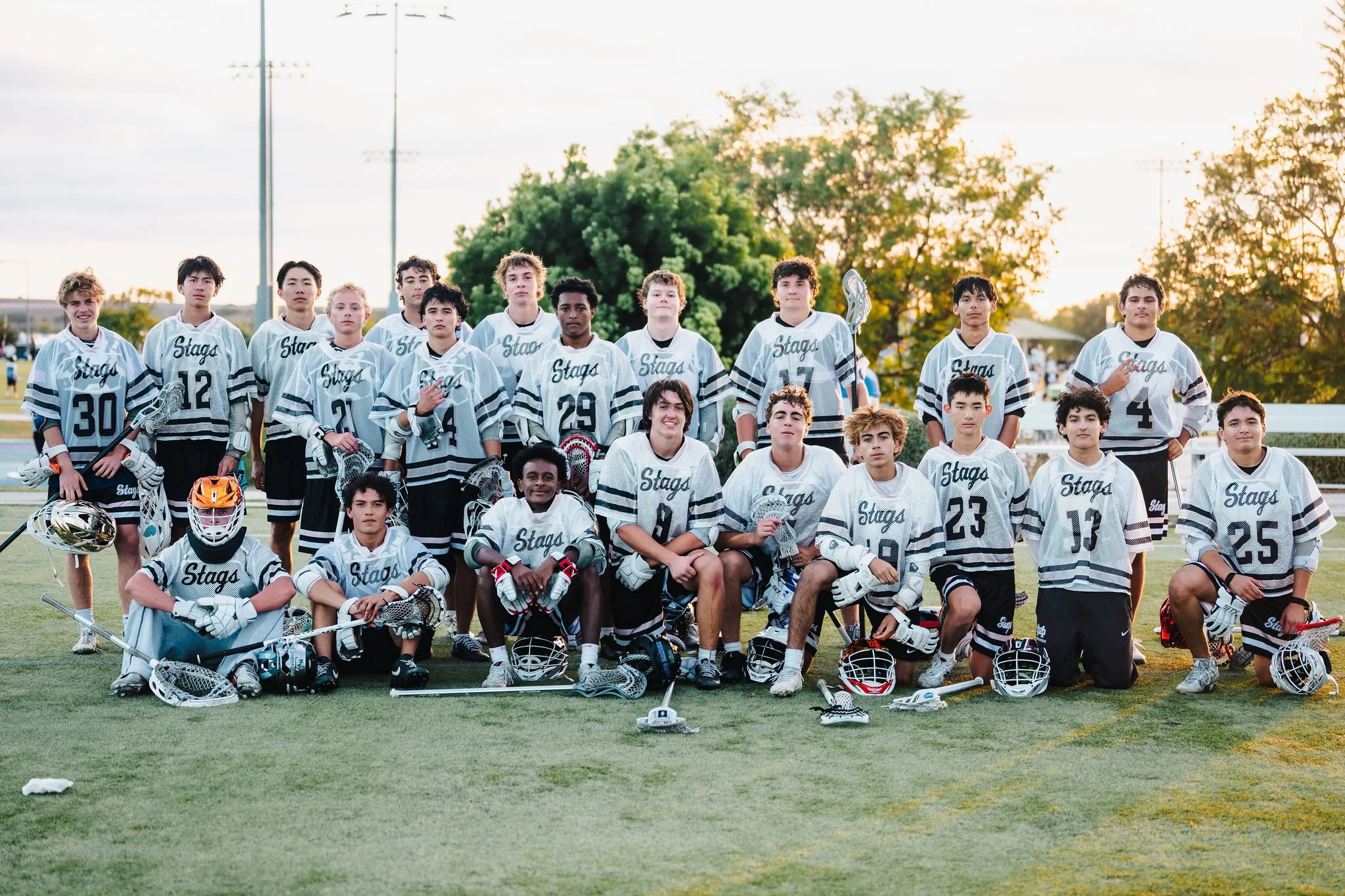 A youth lacrosse team posing for a group photo on a field during sunset. The team members are in uniform, some holding lacrosse sticks and wearing protective gear. There are trees and a sunset in the background.