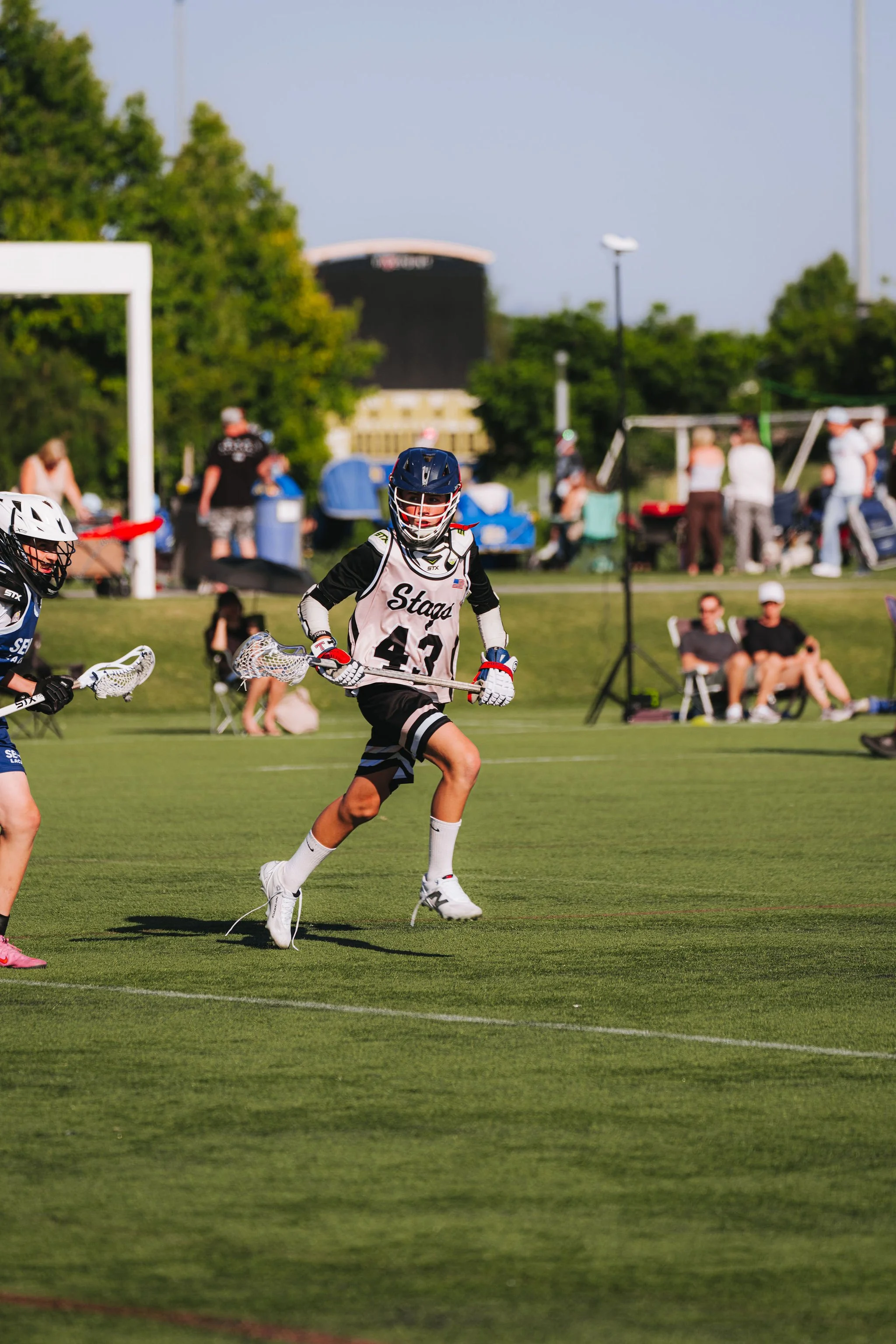 Lacrosse players in black and white uniforms playing on a grass field, with spectators and trees in the background.