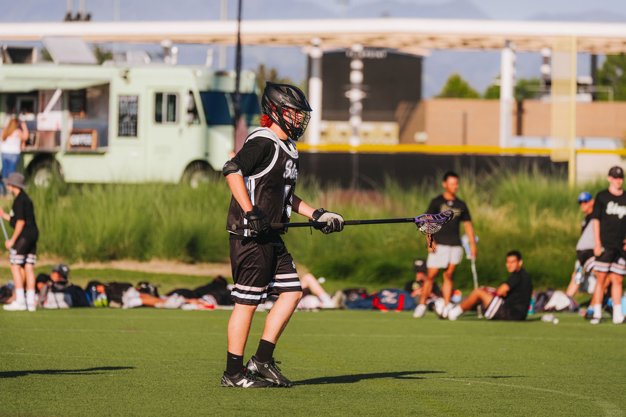 A young person with red hair, wearing a helmet and sports gear, holding a lacrosse stick on a grassy field during a game or practice. In the background, other athletes and a food truck are visible.