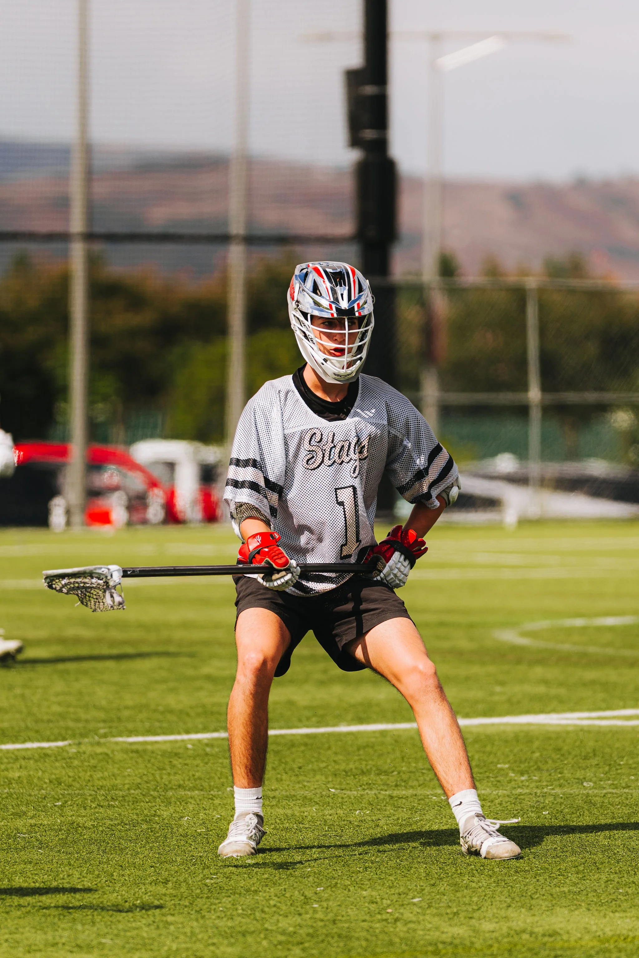 A lacrosse player in a white jersey with black accents, wearing a lacrosse helmet and gloves, standing on a grassy field holding a lacrosse stick, with a chain-link fence and some blurred background structures.