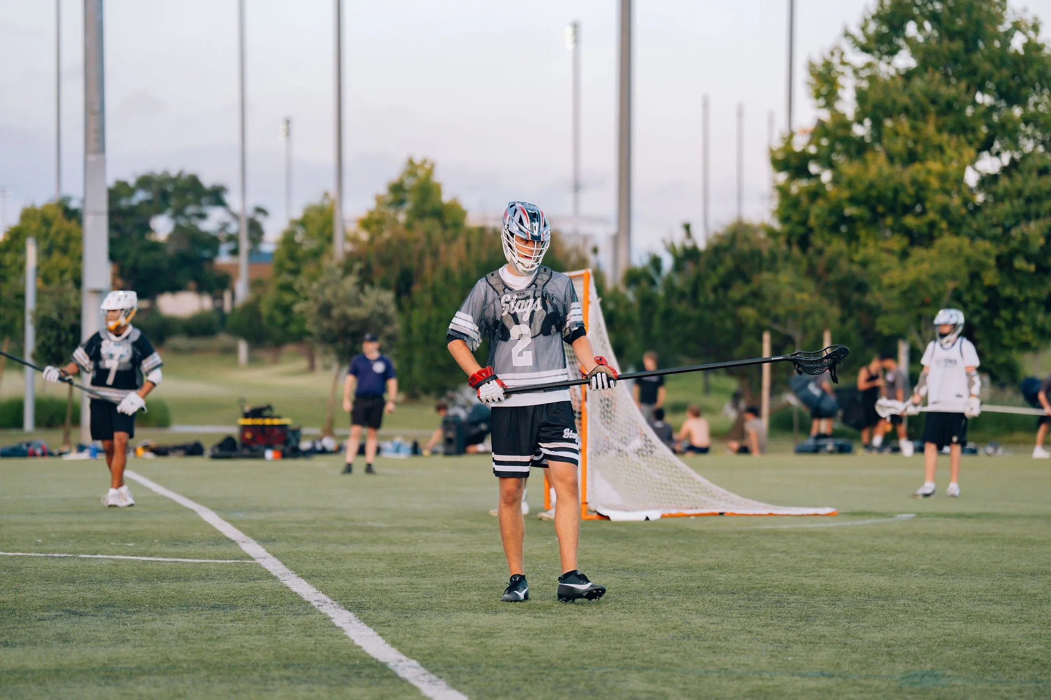 Lacrosse players wearing helmets and jerseys practicing on a field with goal net, trees, and light poles in the background during daytime.