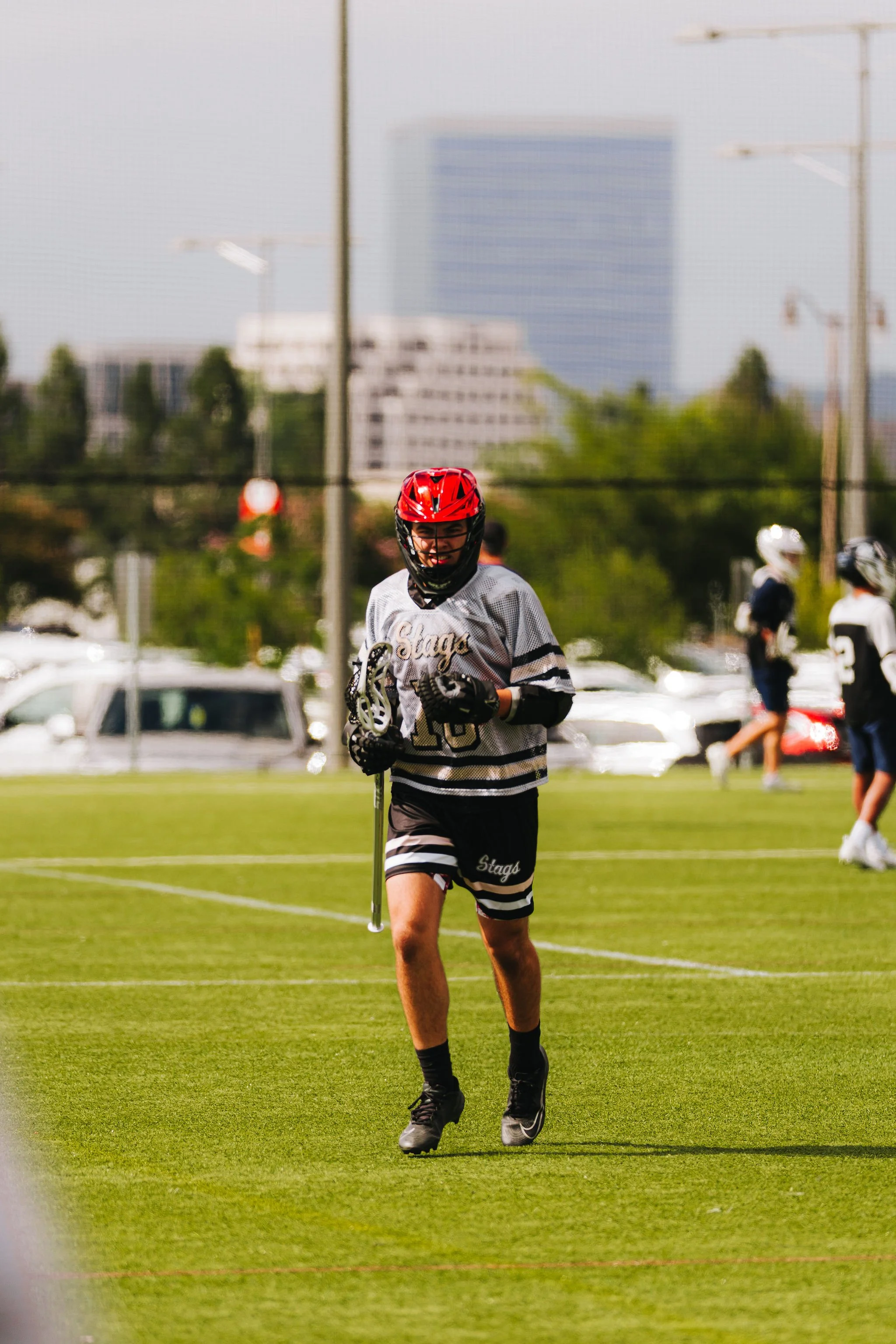 A young male lacrosse player wearing a red helmet and black gloves on a green field, smiling and holding a lacrosse stick, during daylight. Other players and cars are visible in the background.