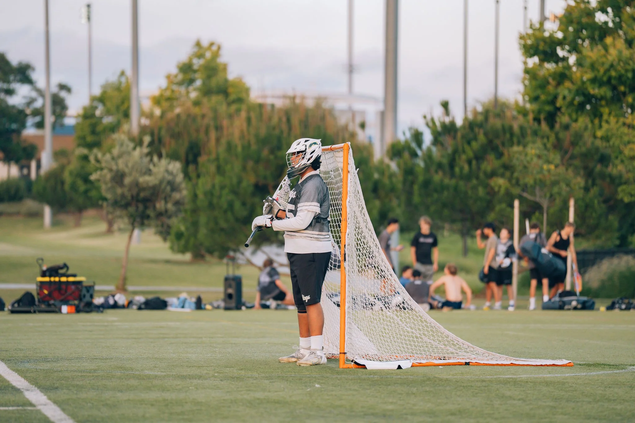 A young lacrosse goalie standing in front of the goalpost on a sports field, wearing a helmet, gloves, and protective gear, with a crowd and trees in the background.