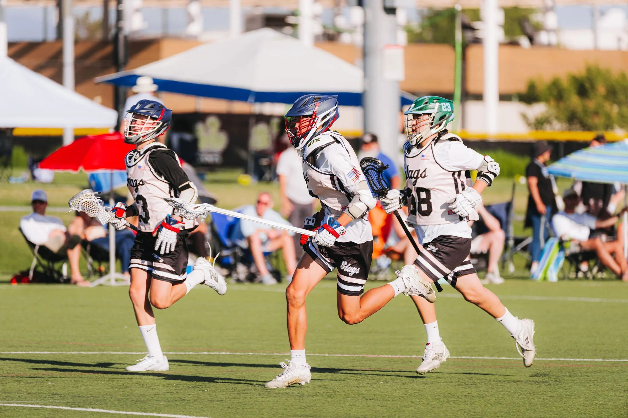 Lacrosse players in black and white uniforms running on a field, wearing helmets and carrying lacrosse sticks, with spectators and umbrellas in the background.
