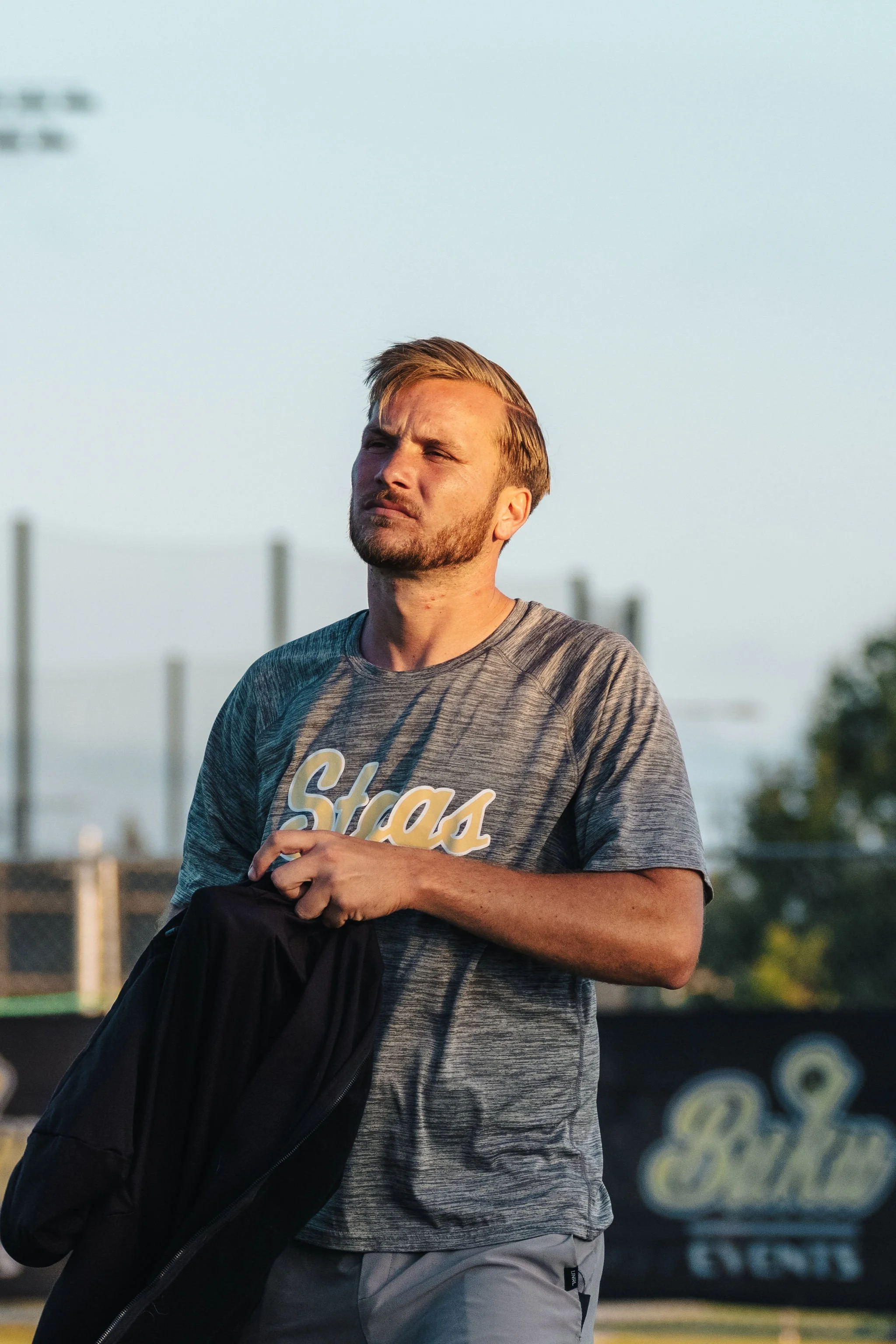 A man with short blond hair and a beard is standing outdoors in the late afternoon, wearing a gray T-shirt with the word 'Stas' printed on it. He is holding a black jacket in his right hand and appears to be looking into the distance with a thoughtfu