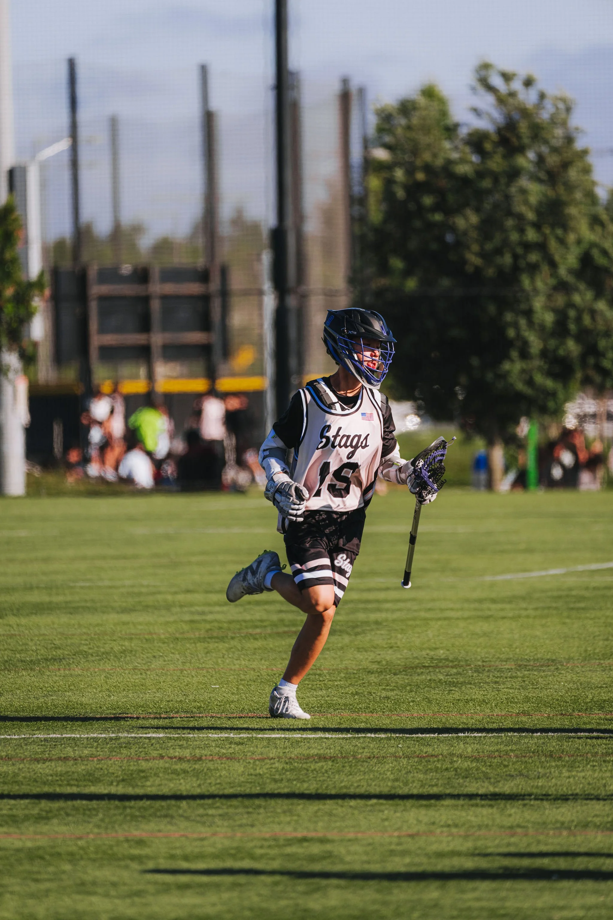 A young lacrosse player running on a field, wearing a helmet and uniform with the word 'Stags' and the number 6, holding a lacrosse stick, with other players and a fence in the background.