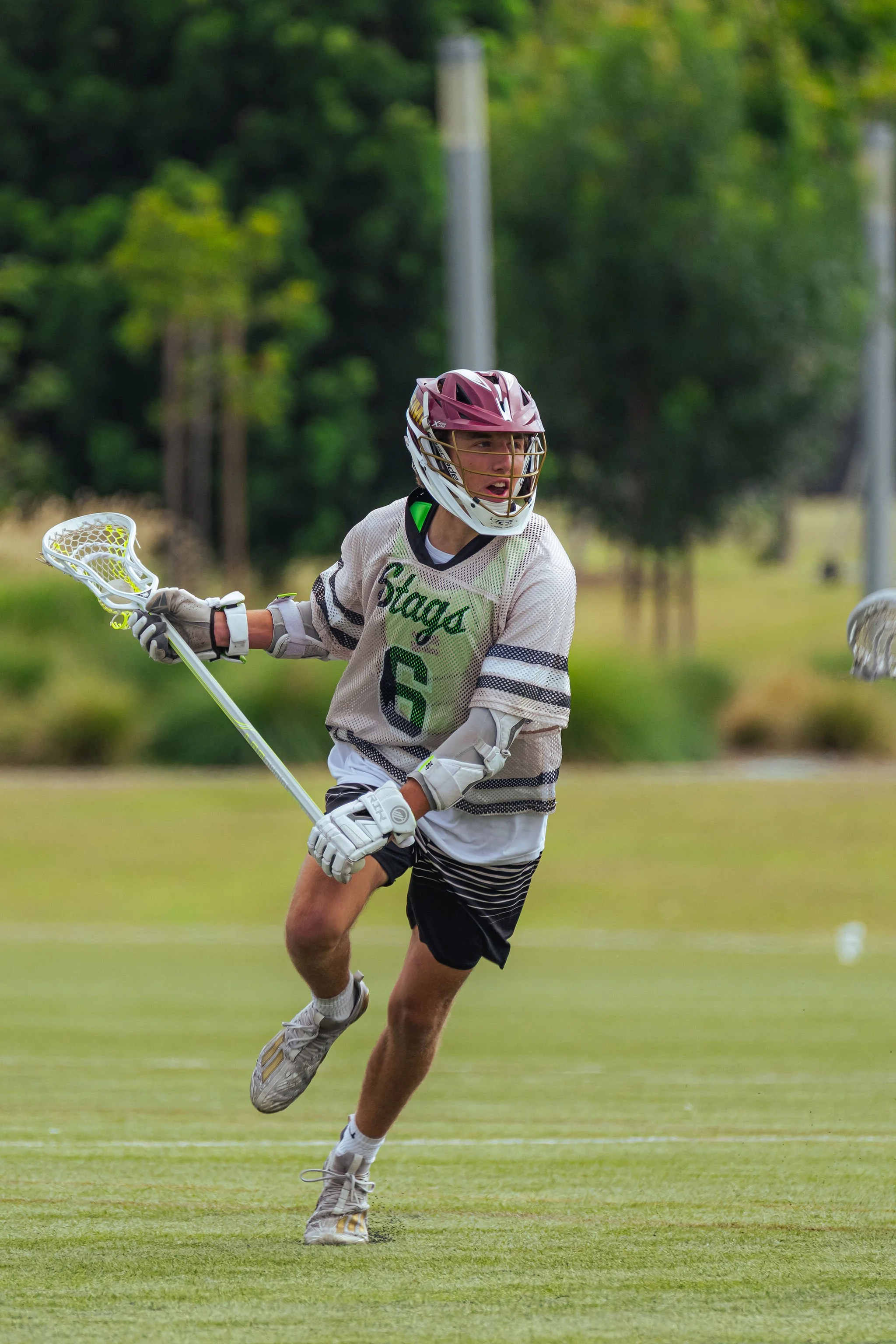 A lacrosse player wearing a maroon helmet, white jersey with green lettering and black shorts, running on a grassy field with trees in the background.