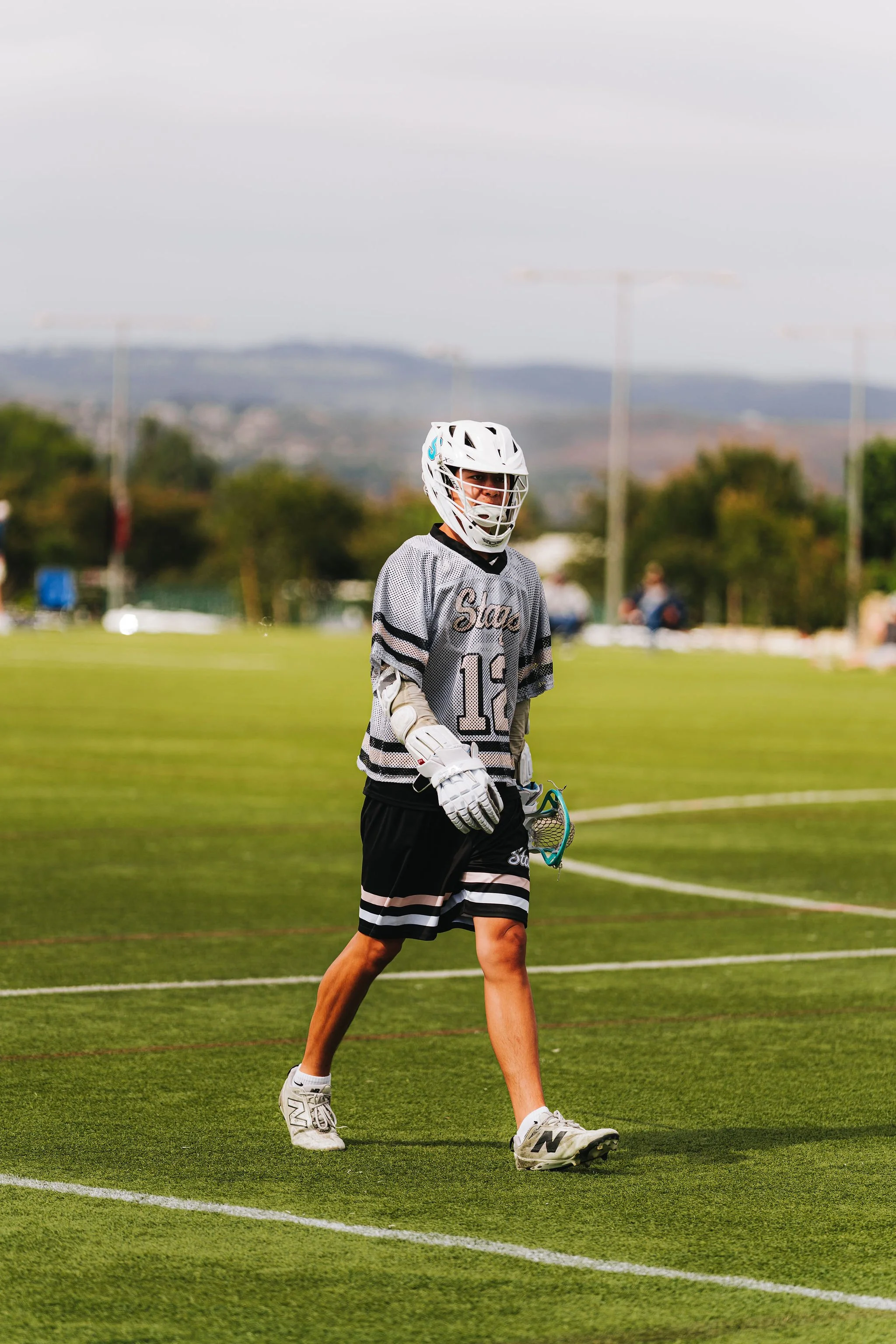 A lacrosse player wearing a white helmet with a face guard, a gray jersey with the word "Stay" and the number 13, black shorts, and white sneakers, standing on a green field with mountains and a cloudy sky in the background.