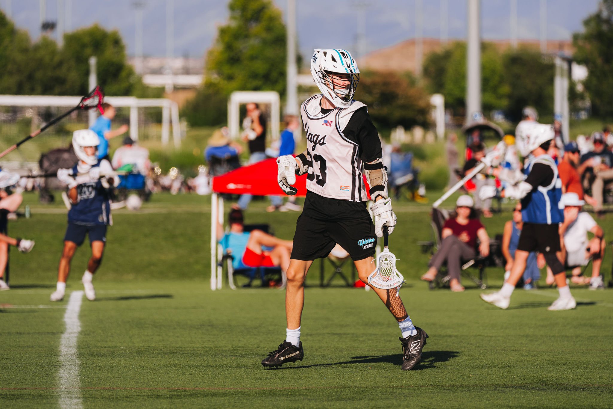 A lacrosse player in black shorts, a white jersey, and a white helmet standing on a green field during a game or practice. Other players and spectators are visible in the background.