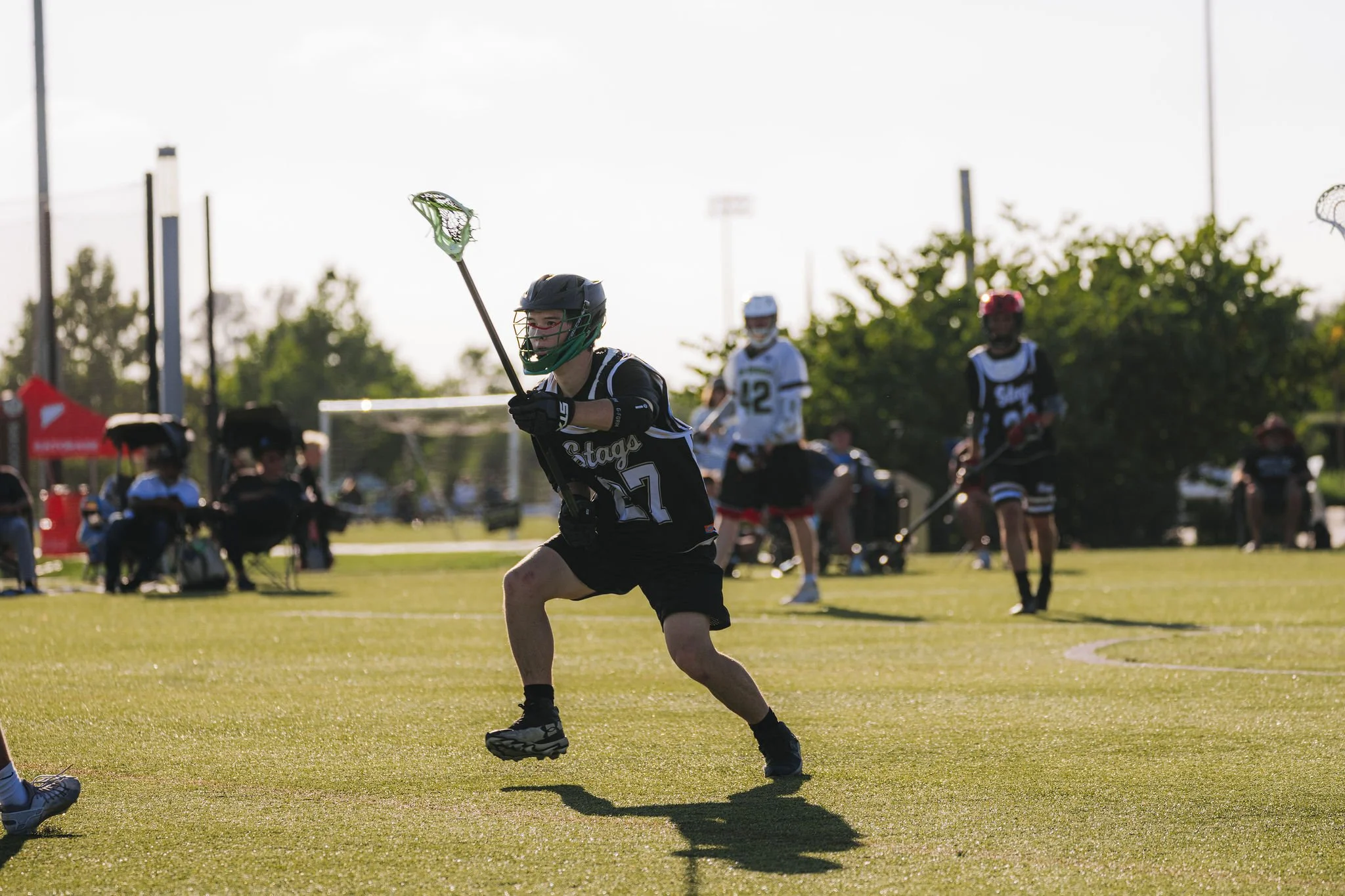 Lacrosse player in black uniform running on grassy field, holding lacrosse stick, with other players and spectators in the background during late afternoon.
