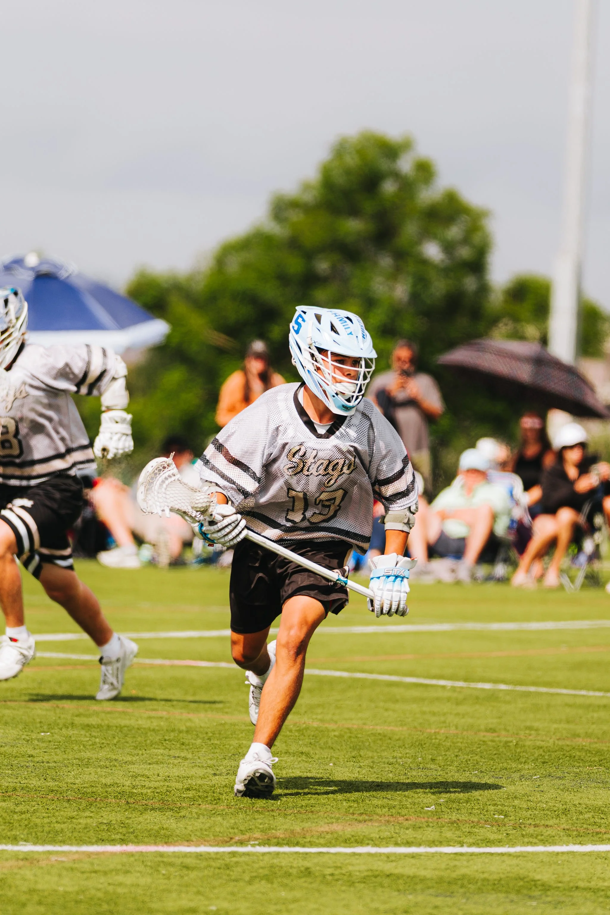 A young male lacrosse player wearing a grey jersey, shorts, a helmet, and gloves, running on a grassy field during a game. Spectators with umbrellas are in the background.