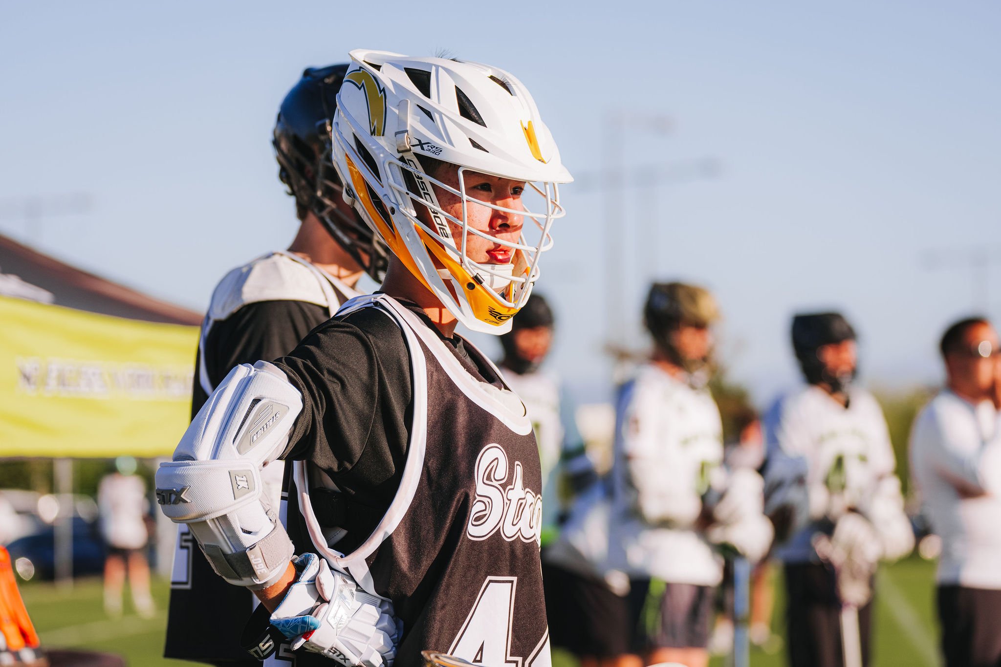 A close-up of a young lacrosse player wearing a white helmet, black and white uniform with the number 4, and protective gear, standing outdoors with other players in the background.