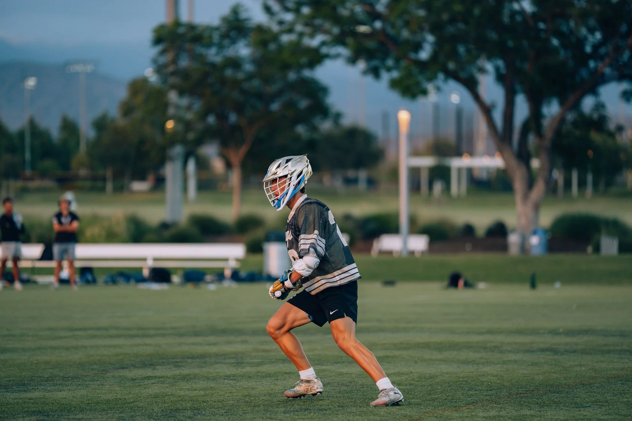 A young female field hockey player in black shorts, a black and white striped jersey, and a white helmet is running on a grassy field during a game in the early evening, with spectators and trees in the background.