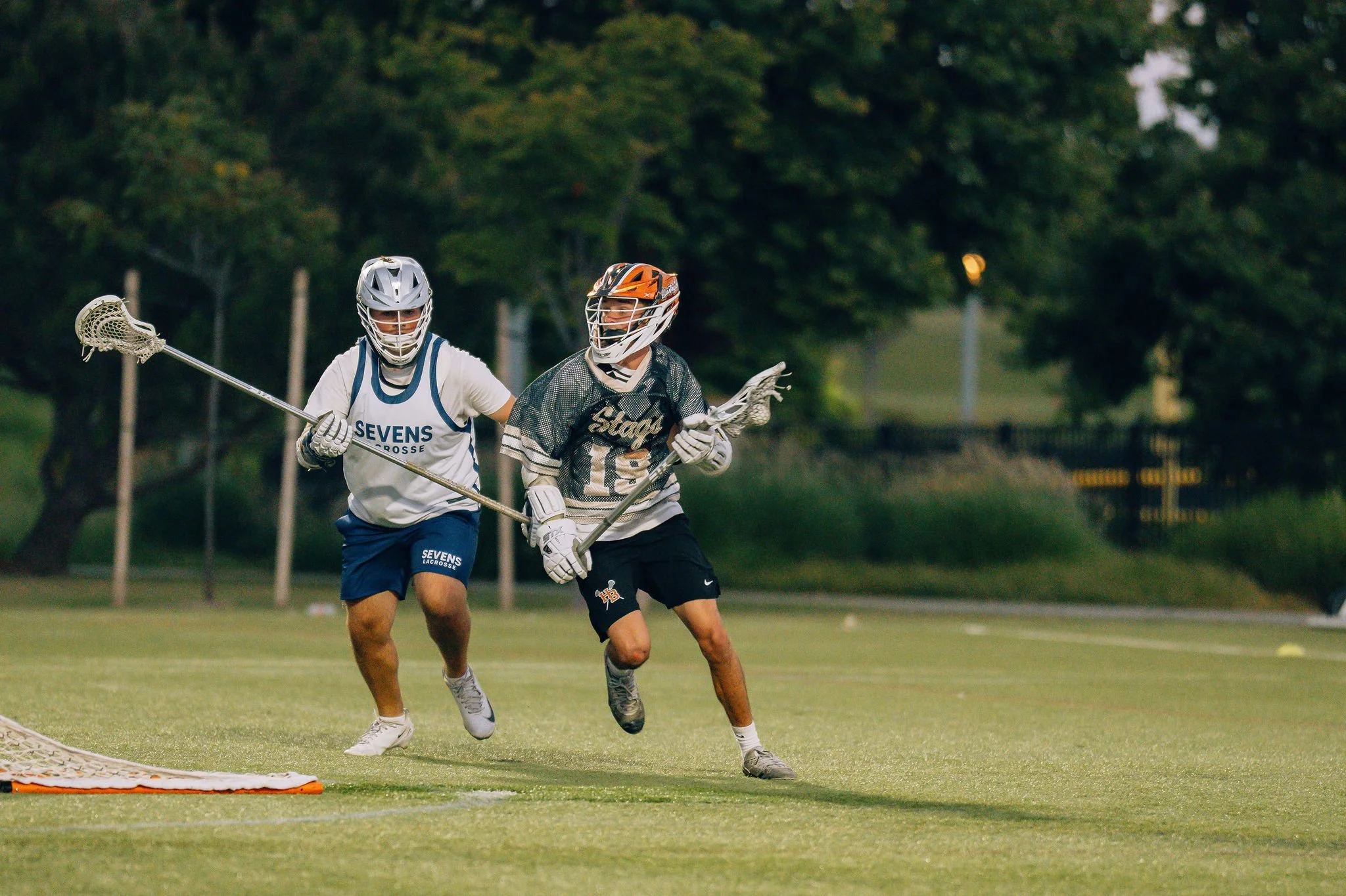 Two athletes playing lacrosse on a field during evening, wearing helmets and protective gear, one in a white jersey with blue shorts and the other in a black and white jersey with black shorts, competing for the ball near a lacrosse goal.