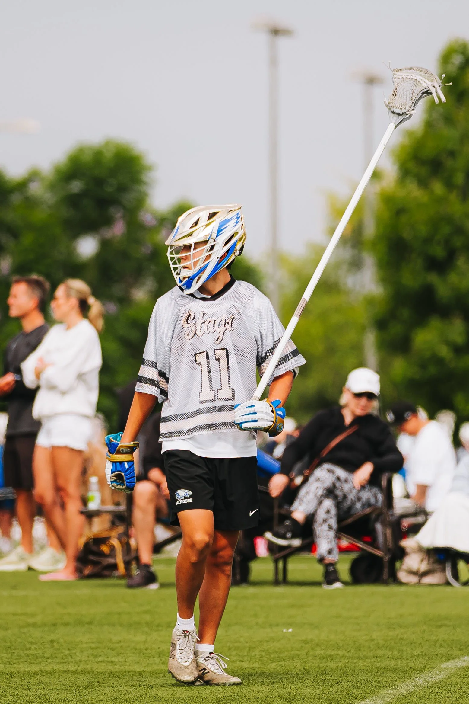 A male lacrosse player in a white jersey with the number 10 and the word 'Stony' on it, wearing a helmet and gloves, holding a lacrosse stick, standing on a grassy field during a game or practice. There are spectators and other people in the backgrou