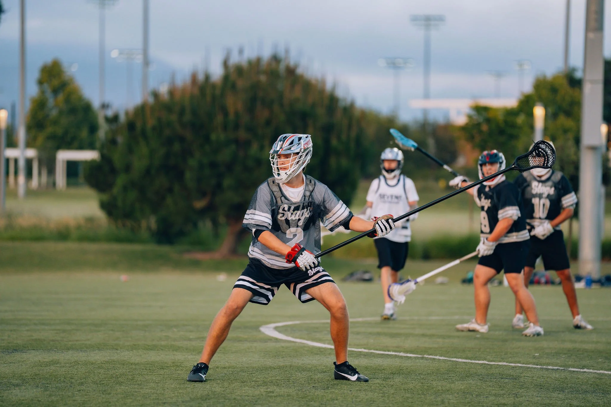 A group of young lacrosse players on a field during daylight, with one player in the foreground preparing to throw a lacrosse ball, wearing a helmet and sports uniform, while others stand in the background.