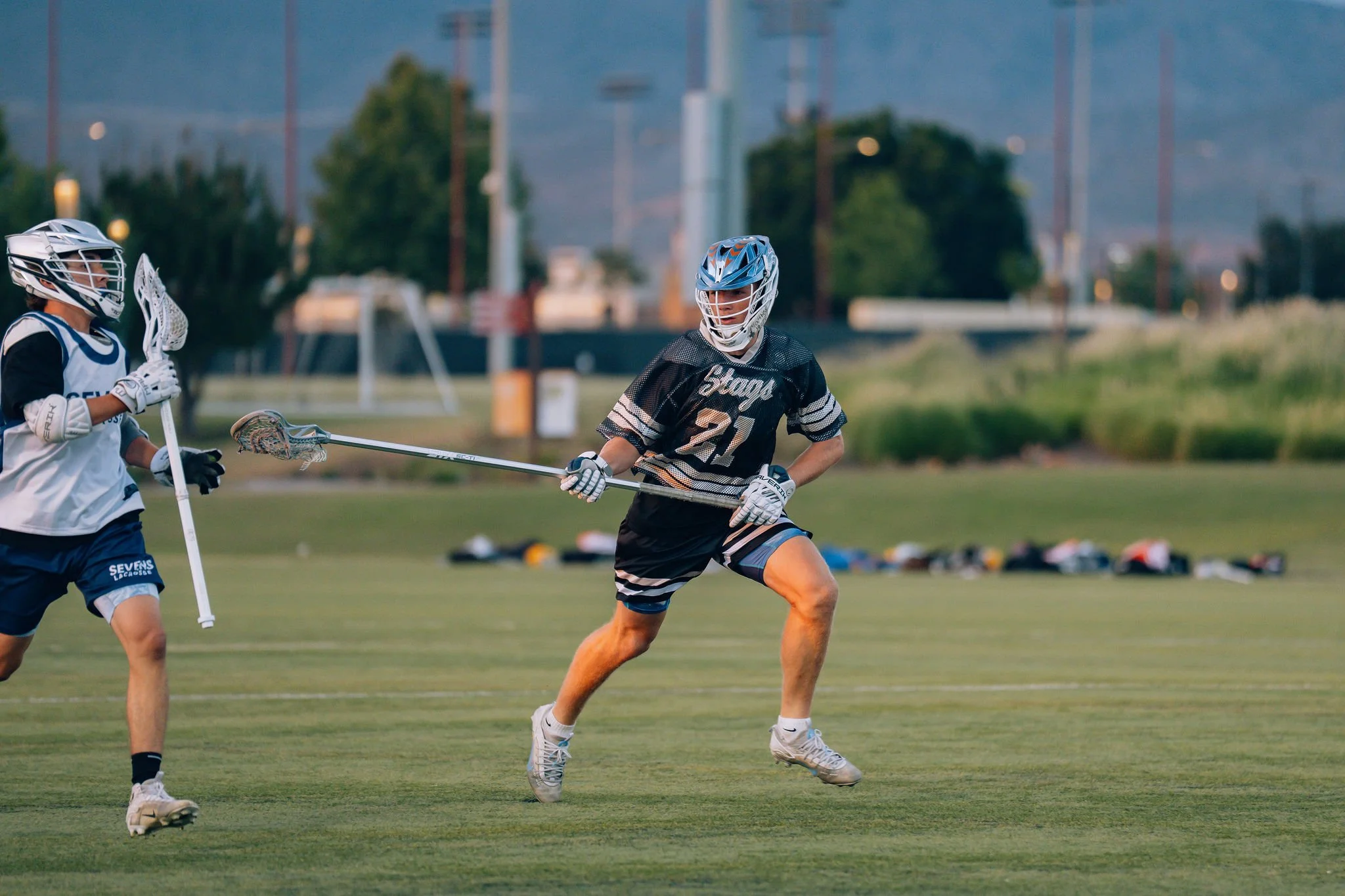 Two young lacrosse players wearing helmets and protective gear, one in a black jersey and the other in a white jersey, compete on a grassy field during dusk.