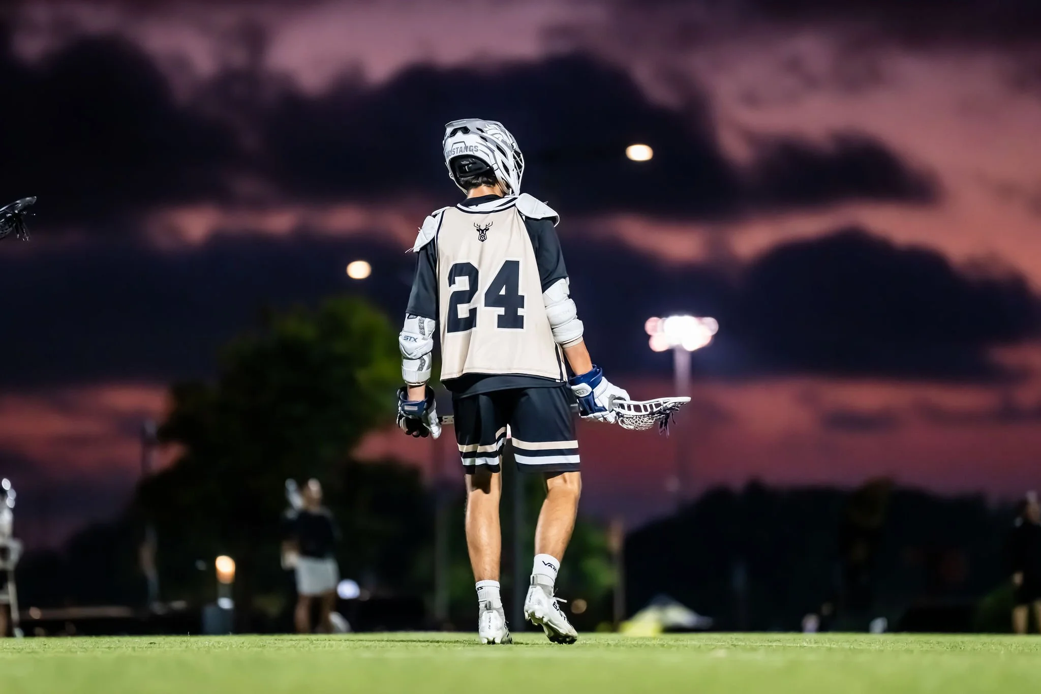 A young ice hockey player wearing a helmet, jersey with the number 24, and hockey gloves, holding a hockey stick, standing on a green field during dusk with a dark cloudy sky and the moon visible.