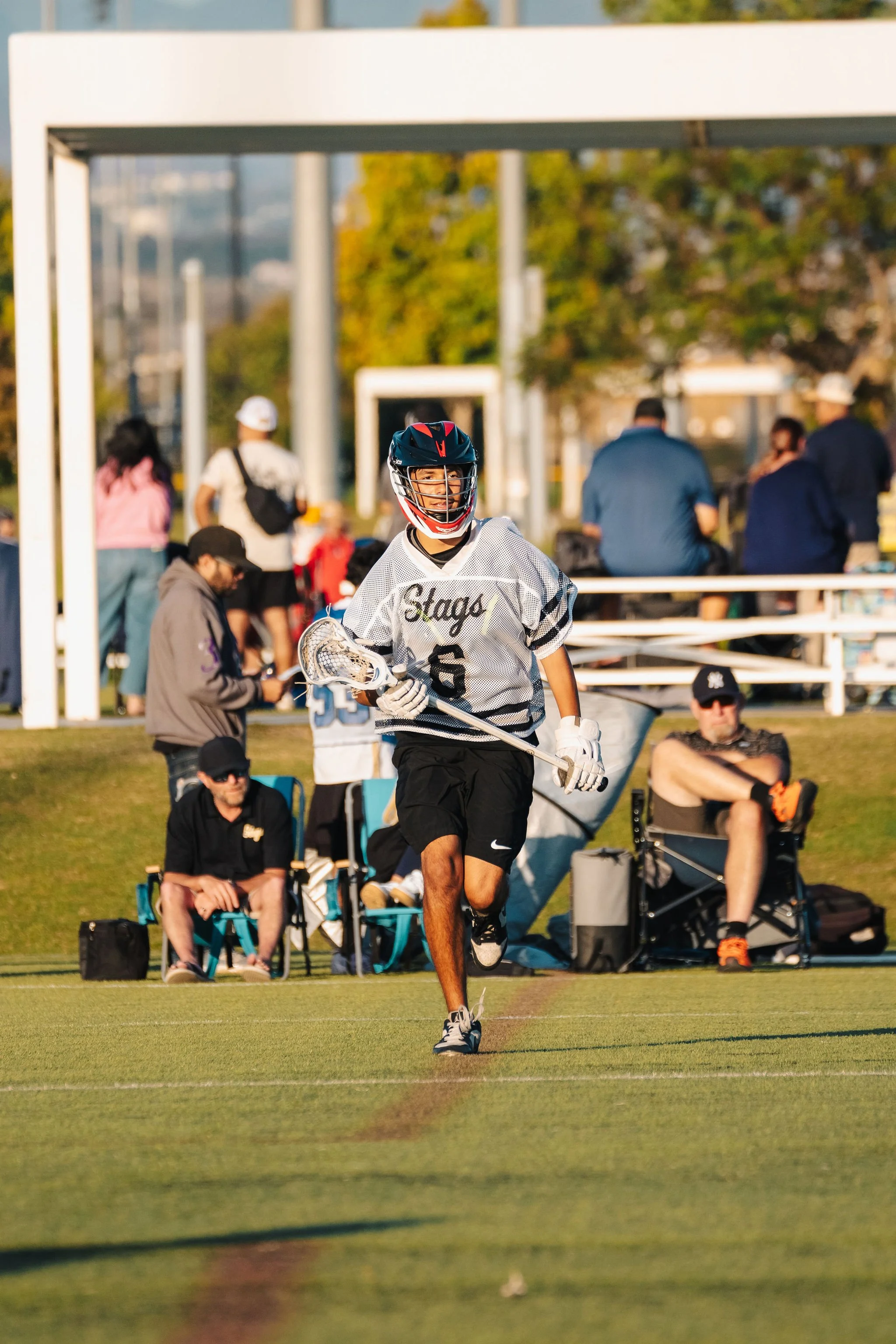 Lacrosse player wearing a helmet and jersey, running on field during game, with spectators and other players in background.