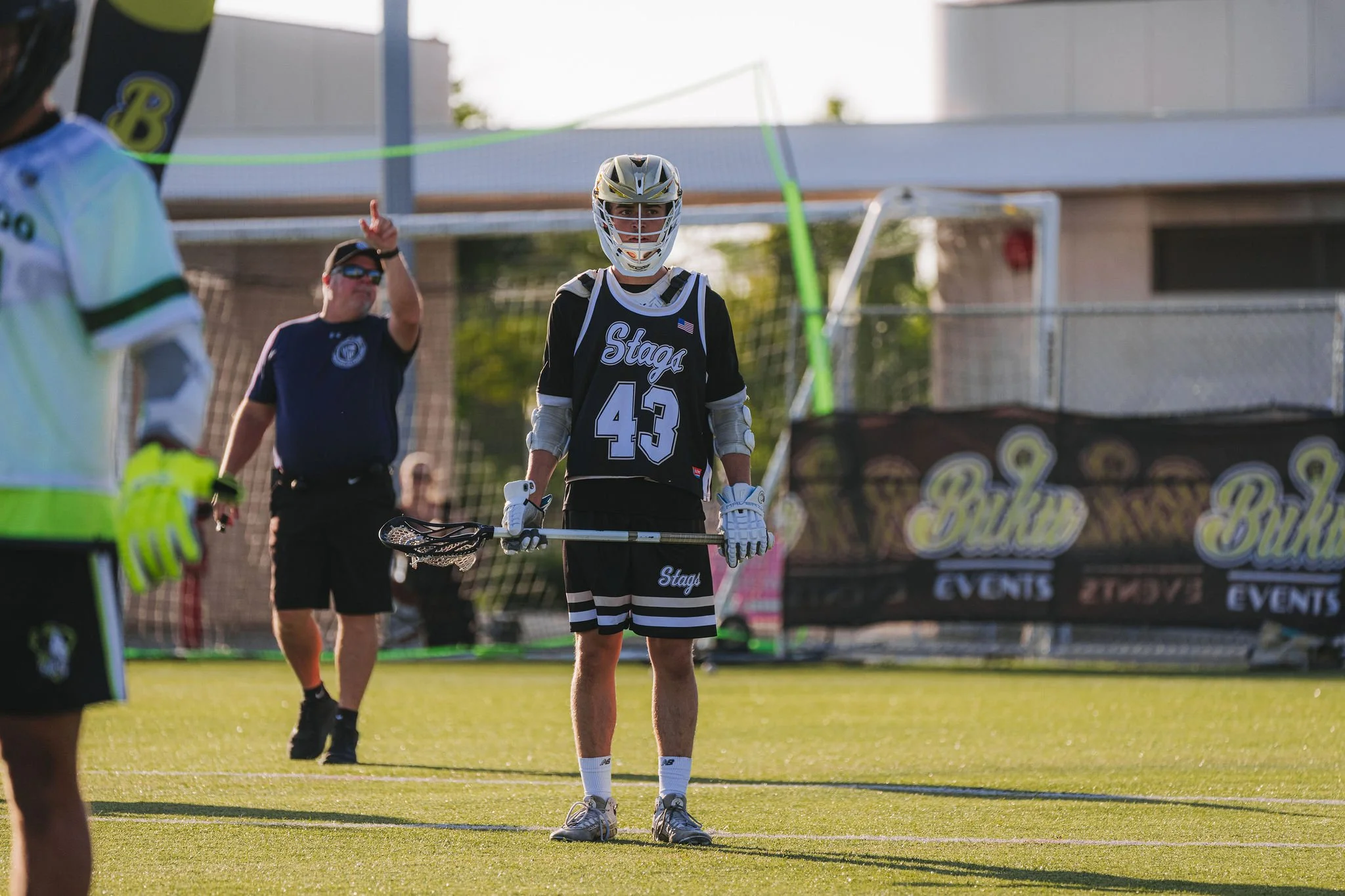 A lacrosse goalie wearing a helmet and jersey with the number 43 stands on the field holding a lacrosse stick, with players and officials nearby during a game.