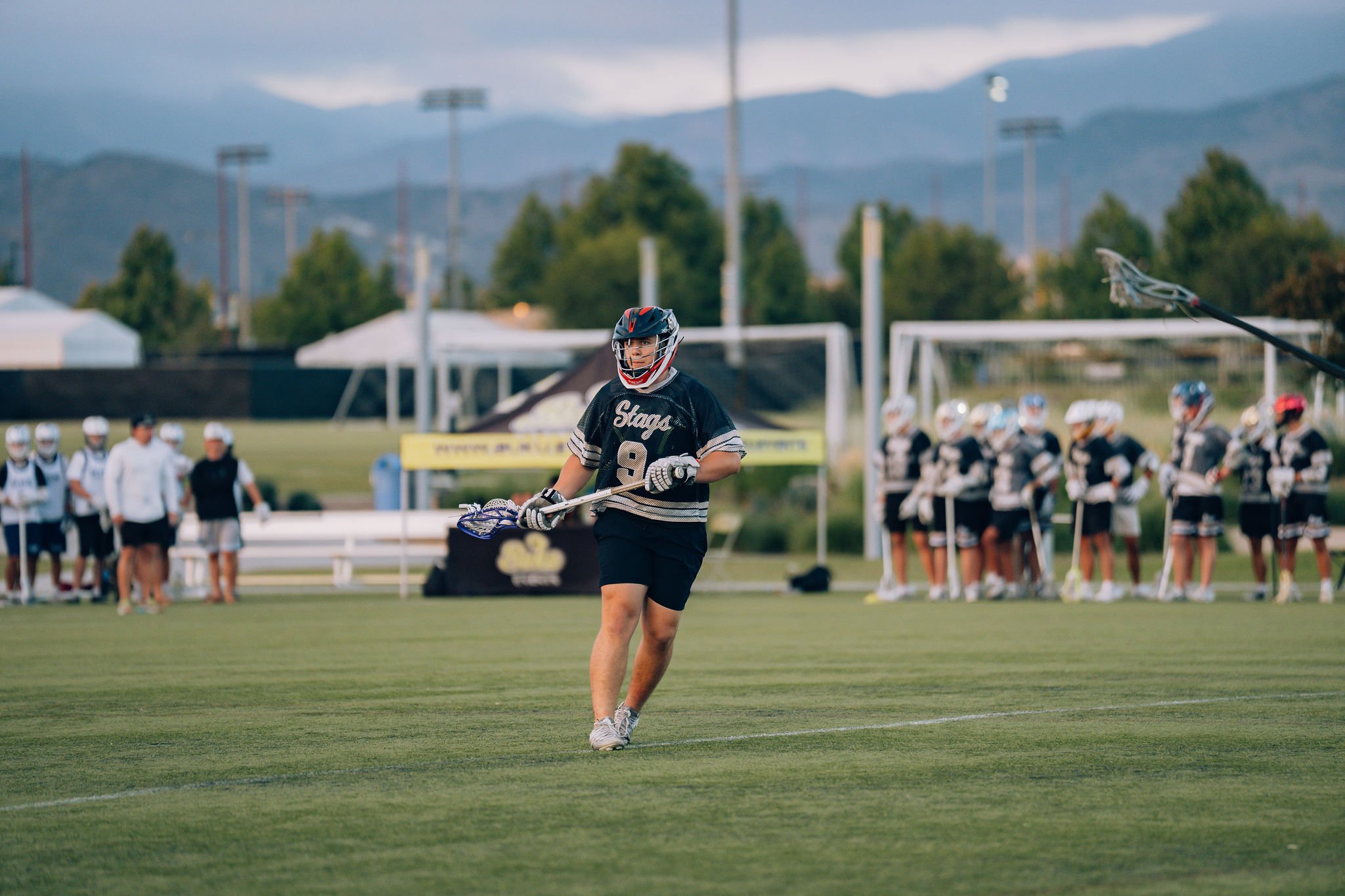 An athlete in a black sports jersey and helmet walking on a lacrosse field, with a group of players and coaches in the background. The scene is outdoors with mountains and cloudy sky in the distance.