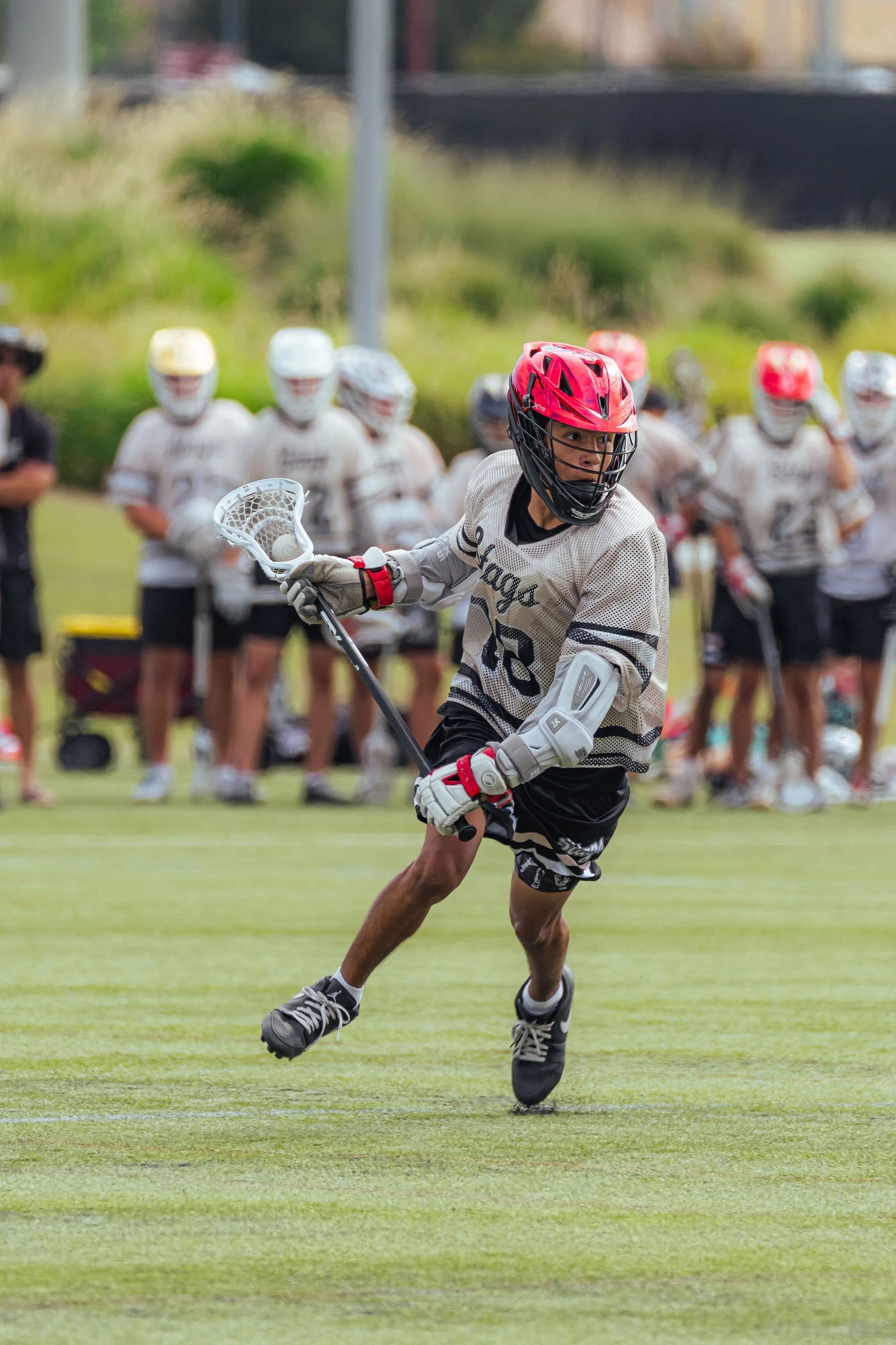 A young male lacrosse player wearing a red helmet, white jersey, and black shorts is running on a field holding a lacrosse stick. In the background, there are several other players in white jerseys and helmets, along with a person in a black hat, on 