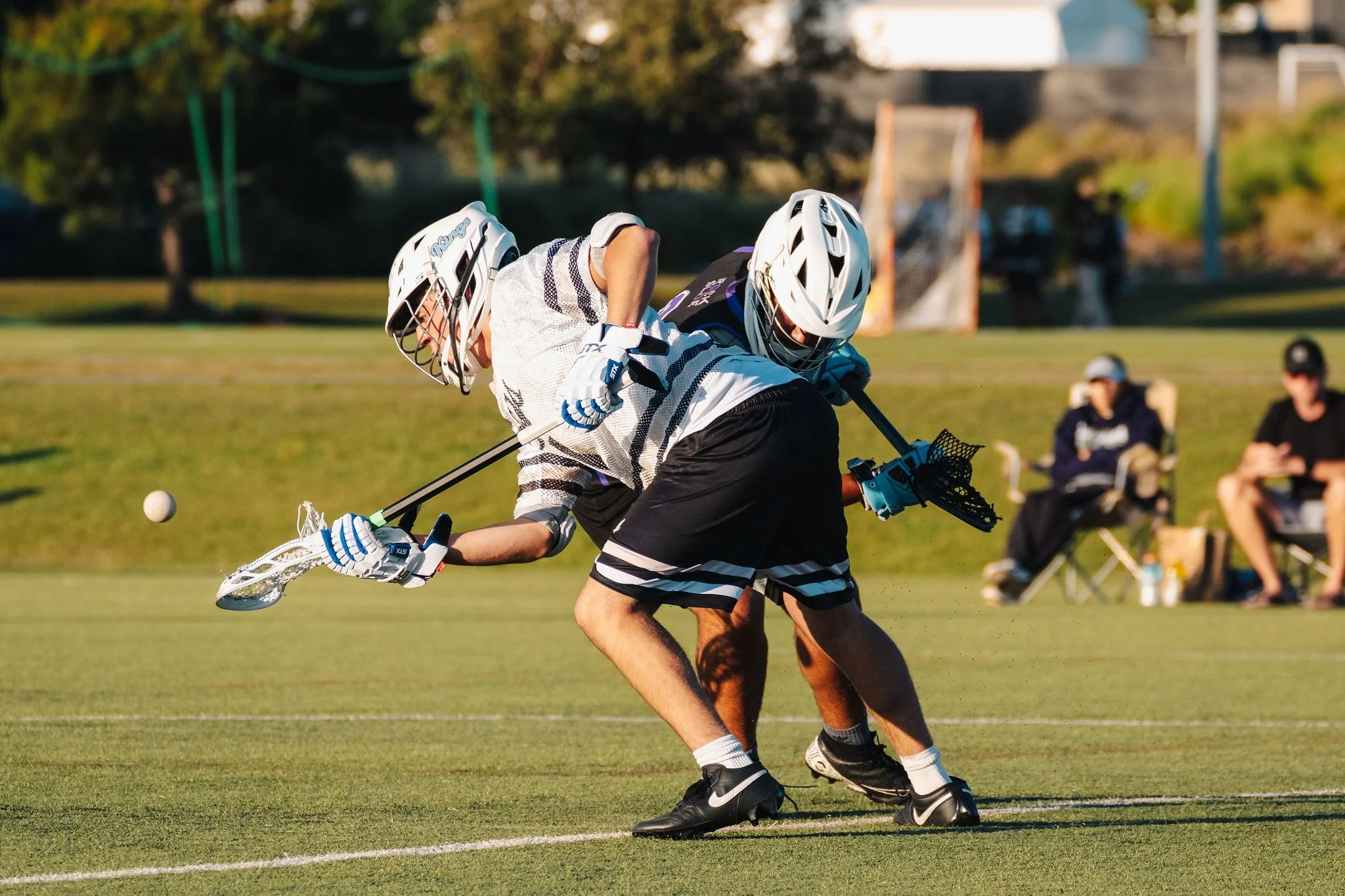 Two lacrosse players in helmets and jerseys compete for the ball on a grassy field, with spectators seated and watching in the background.