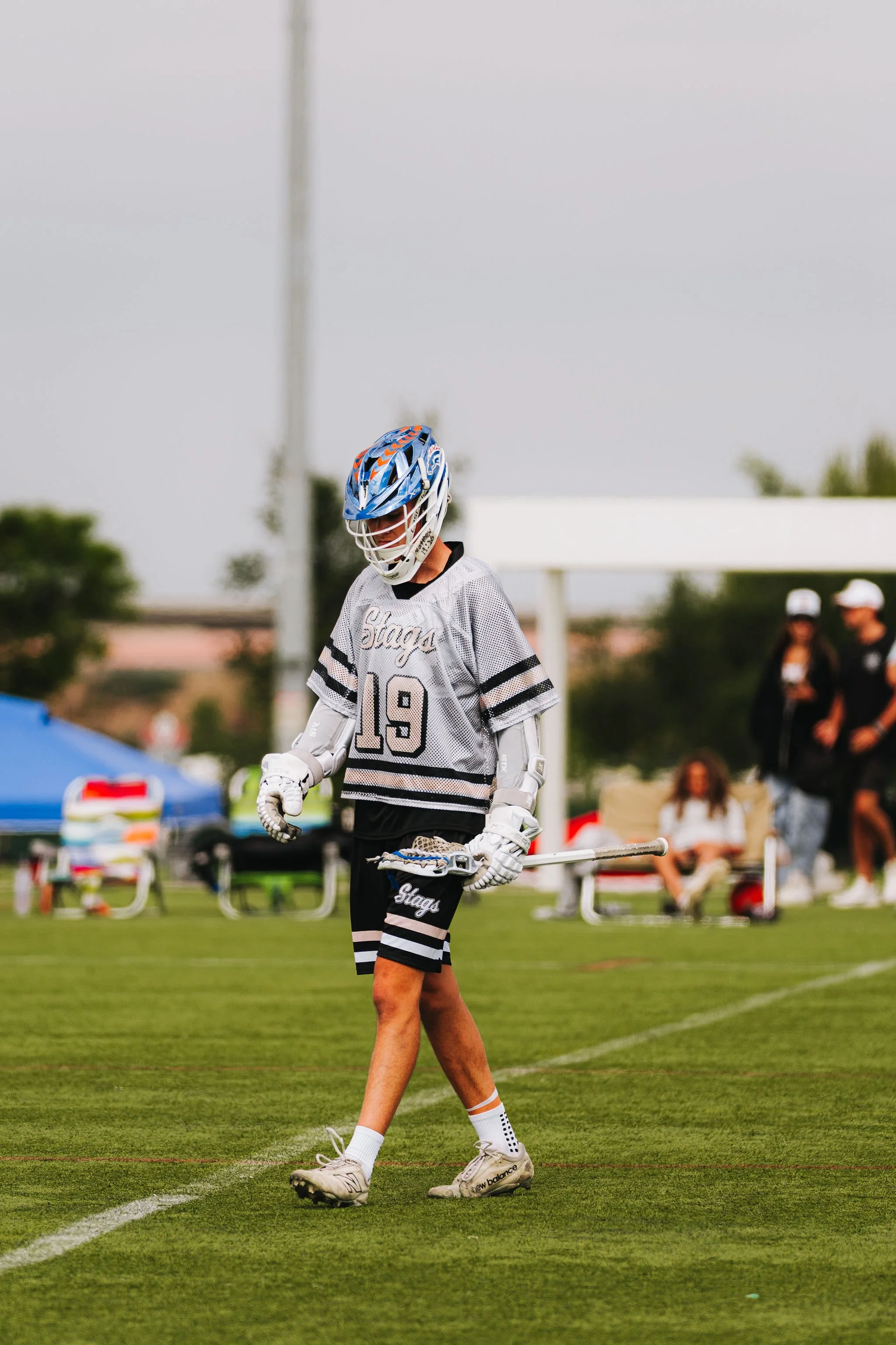 A young male athlete wearing a hockey jersey with the number 19, a helmet, gloves, and sports shoes, walking across a well-maintained grassy field during a game or practice session.
